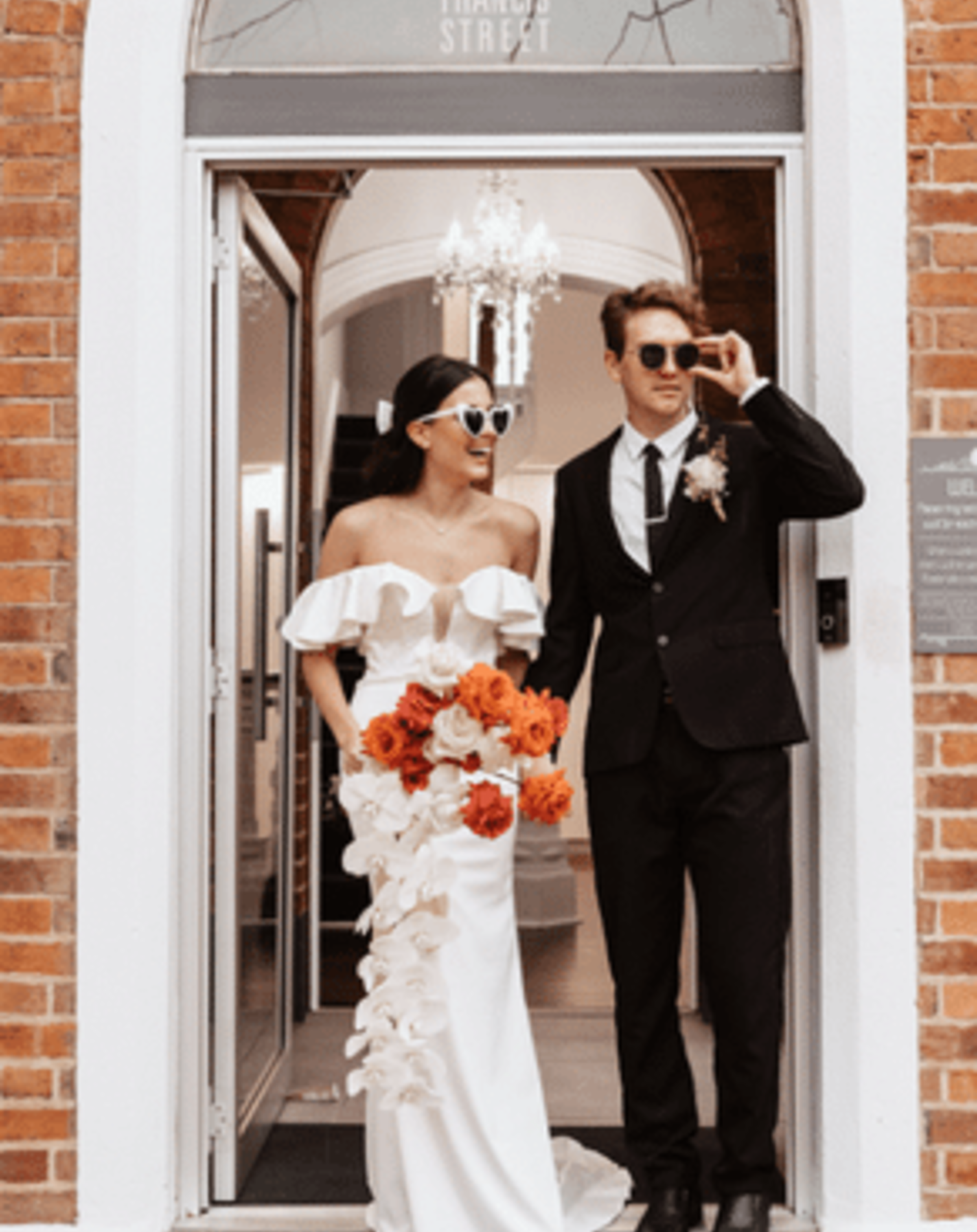 Stylish bride and groom in sunglasses stand in a doorway, with the bride holding a bold orange and white bouquet.
