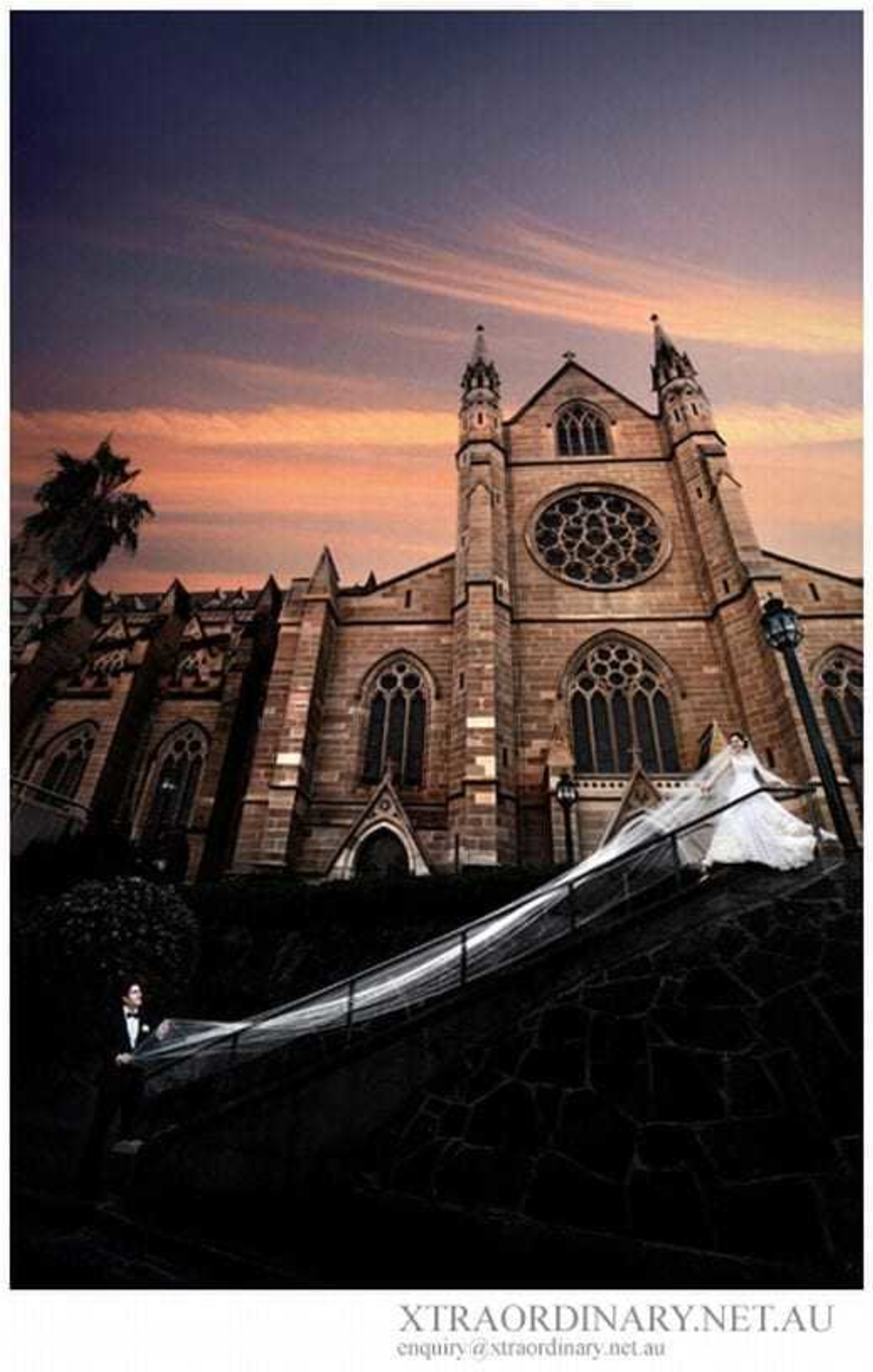 Bride and groom pose outside a grand cathedral at sunset, her long veil flowing dramatically down the stone steps.