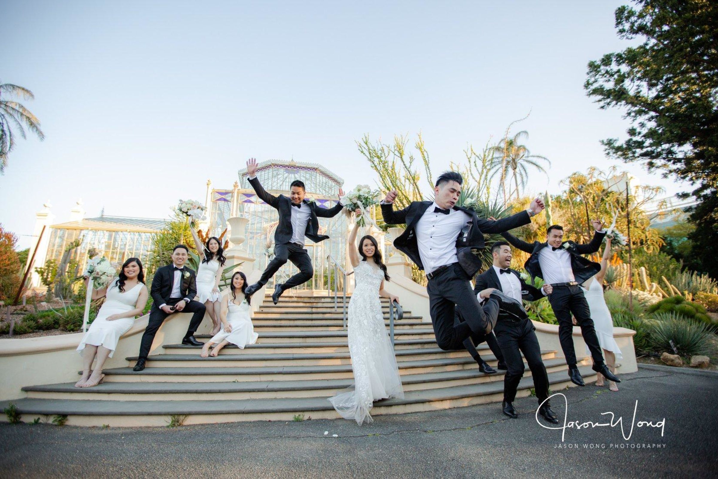Joyful wedding party jumps on grand outdoor steps in front of a glass conservatory.
