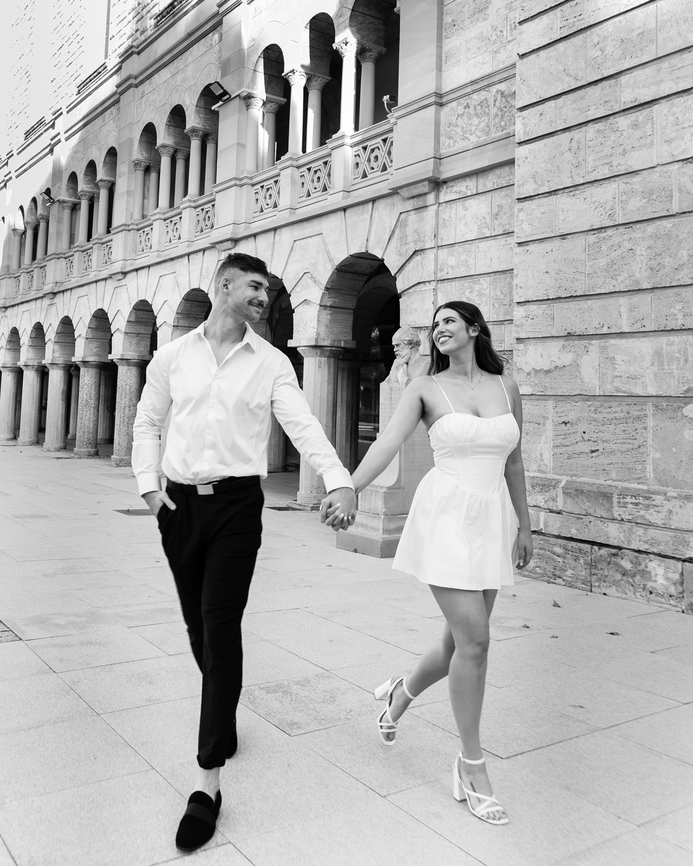 Smiling couple holding hands and walking past a grand stone building in a black and white engagement photo.