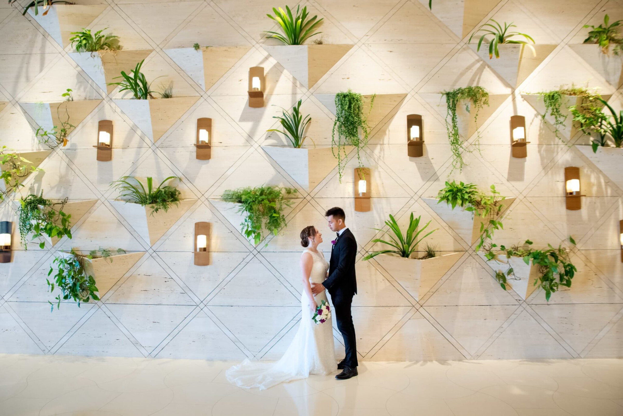 Bride and groom stand facing each other in front of a modern indoor wall with plants and geometric lighting.
