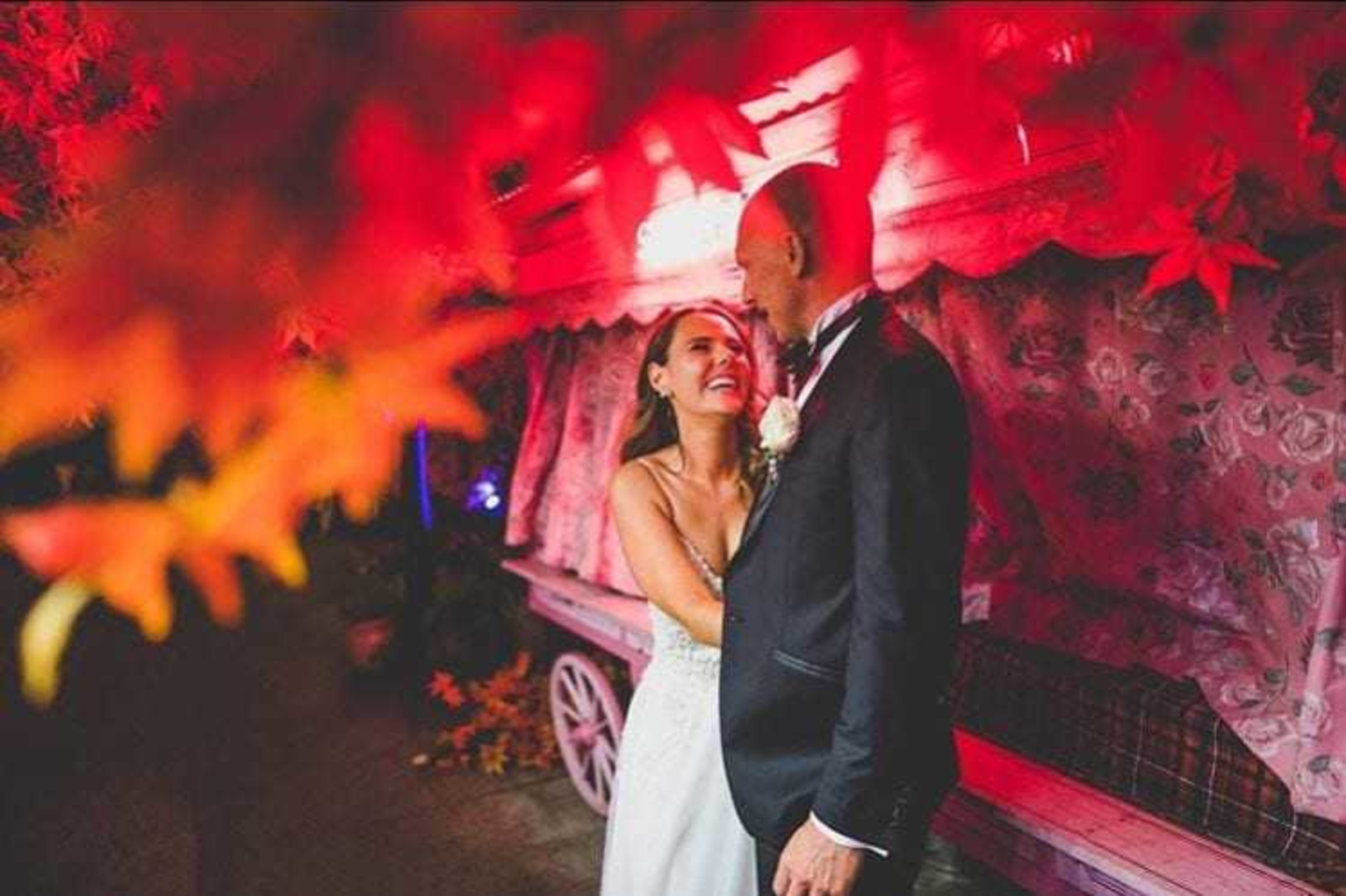 Bride and groom share a joyful moment under vibrant red foliage and colorful evening lighting.
