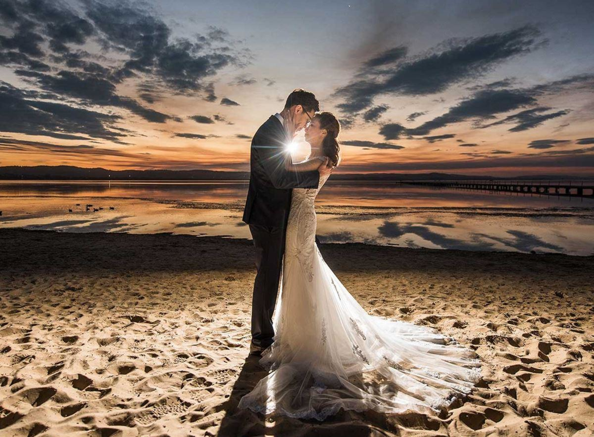 Bride and groom embrace on a sandy beach at sunset with dramatic sky and reflections over the water.