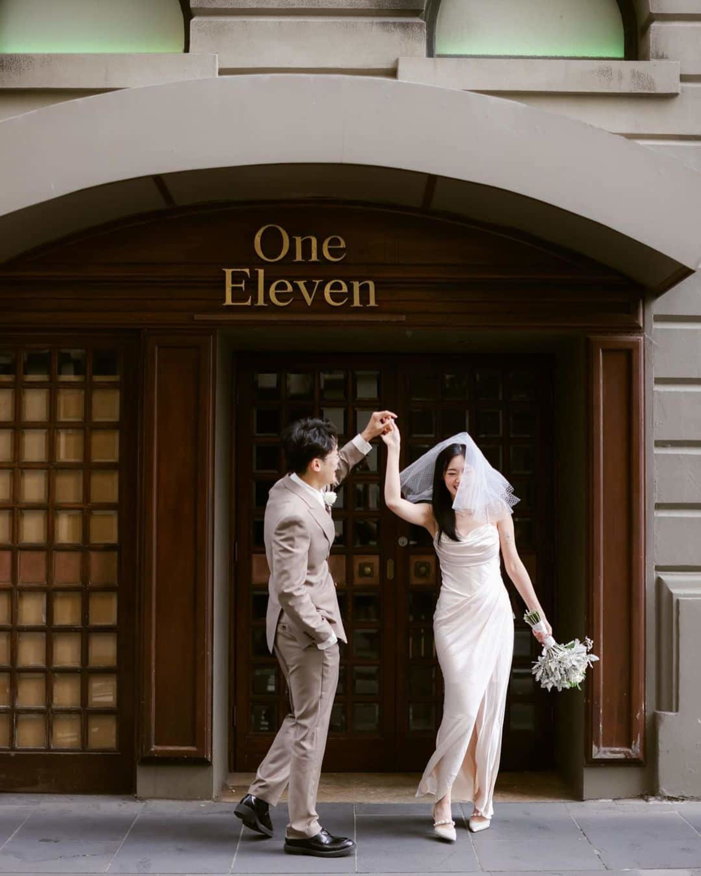 A bride and groom dance happily outside an elegant city venue entrance labeled One Eleven.