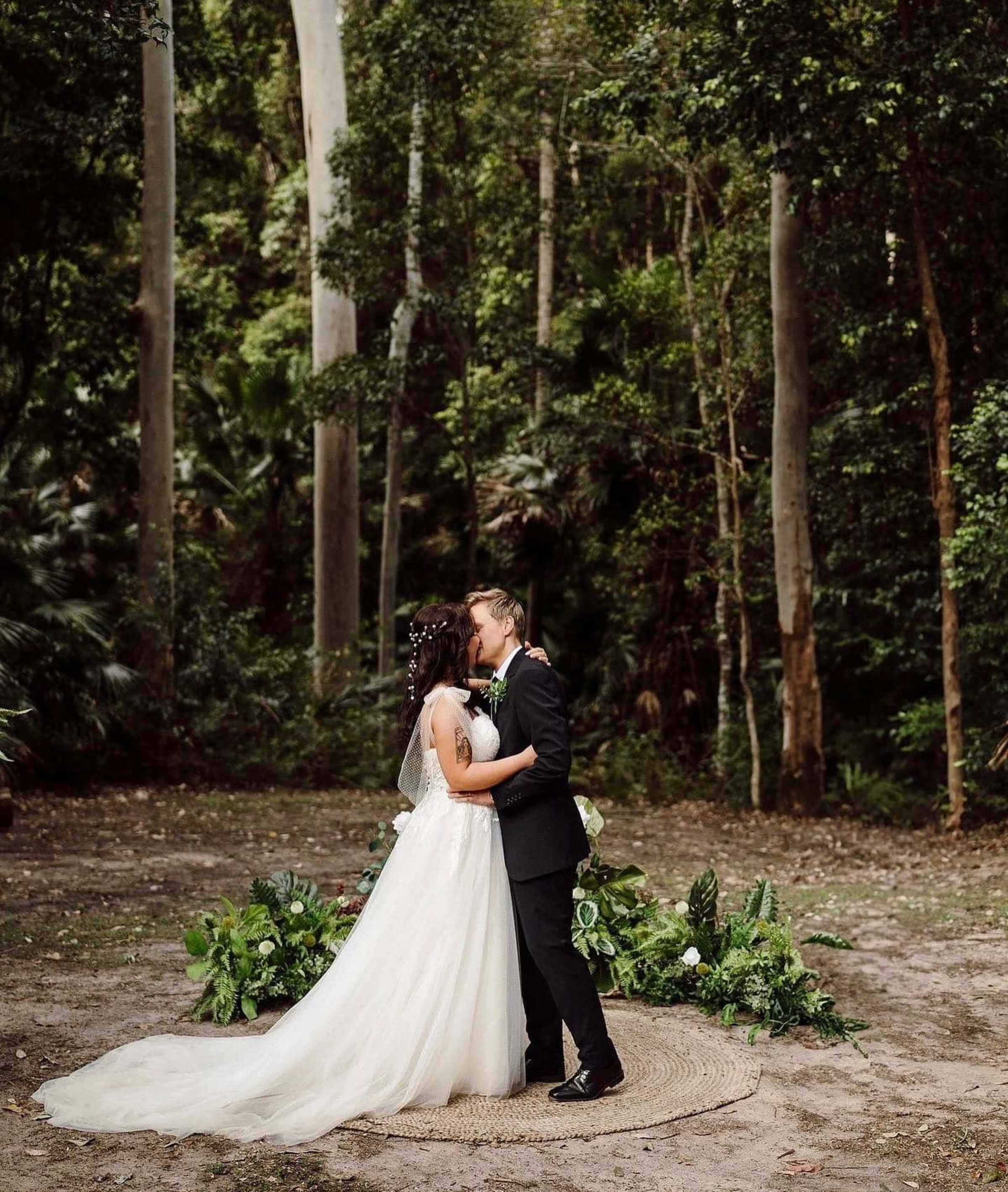 A couple kisses during their forest wedding ceremony on a natural rug surrounded by greenery.