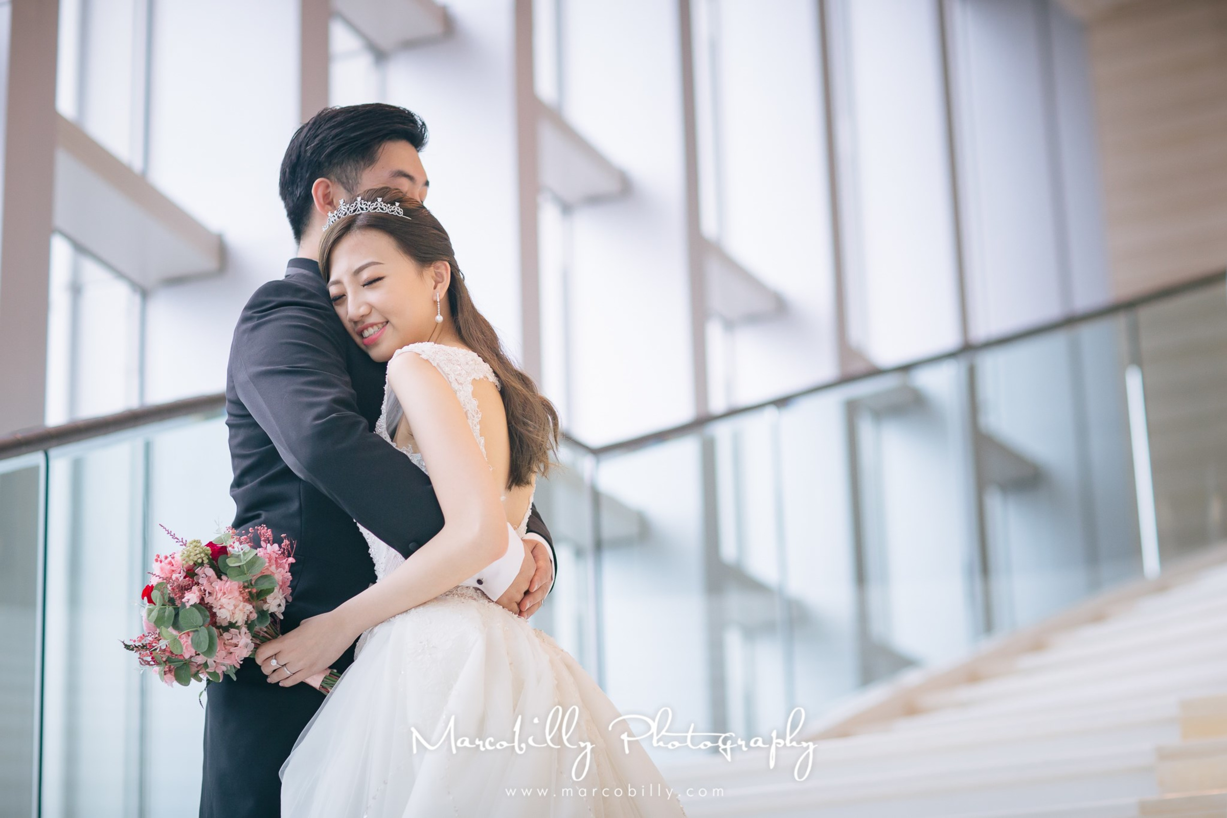 Smiling bride in a white gown hugs her groom on a bright modern staircase, holding a pink and red bouquet.