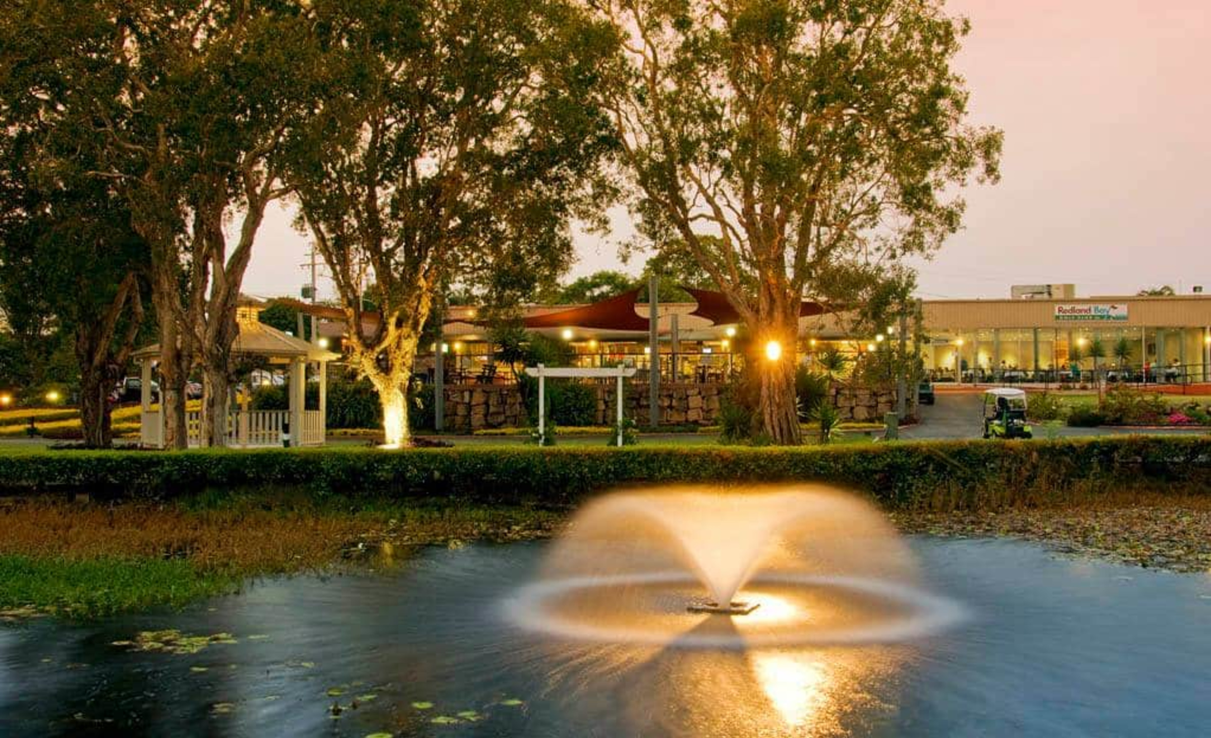 Evening view of a garden wedding venue with gazebo, illuminated trees, and a fountain on a tranquil pond.