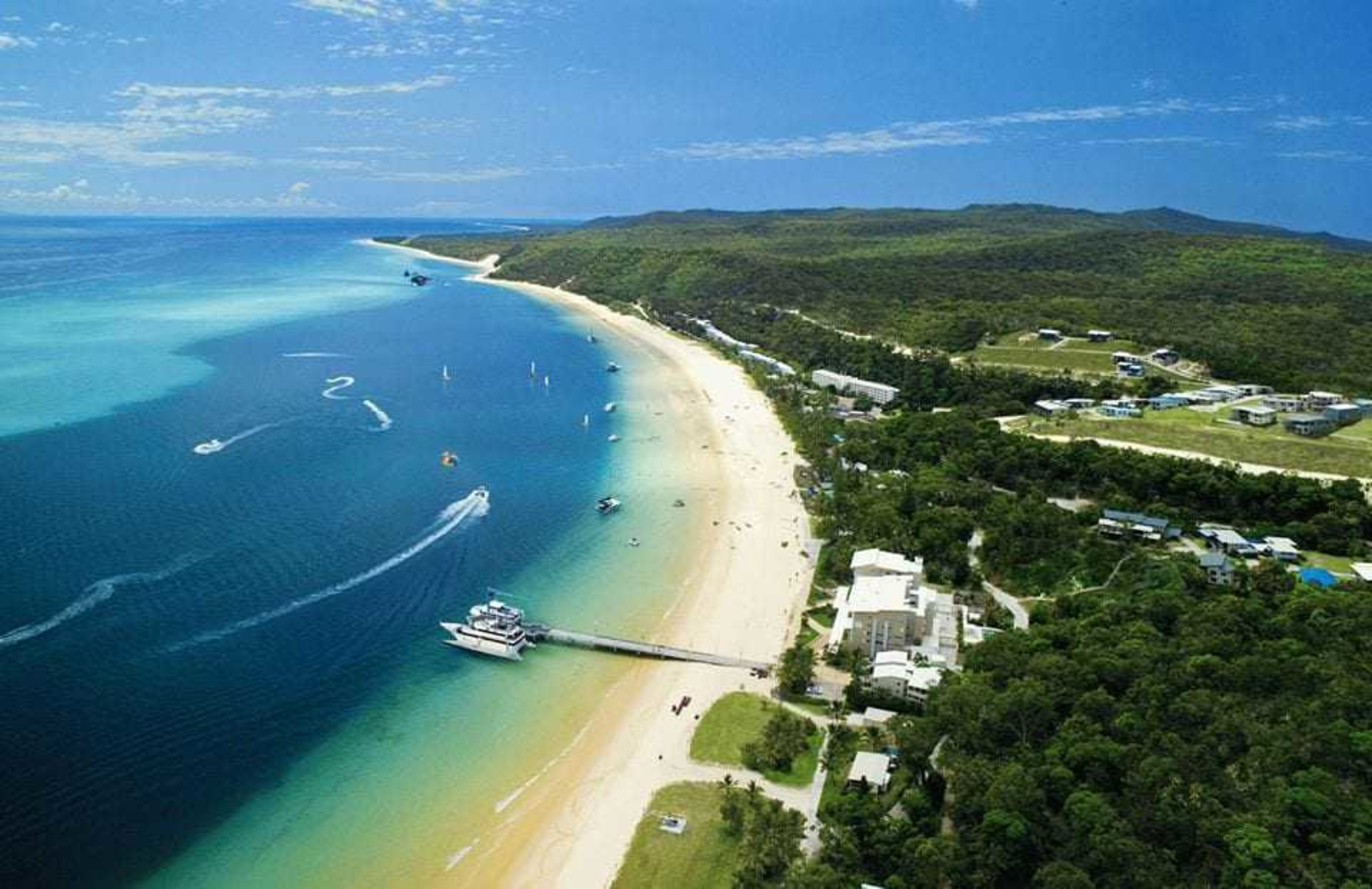 Aerial view of a tropical beach resort with a long pier, turquoise water, and lush green coastline.
