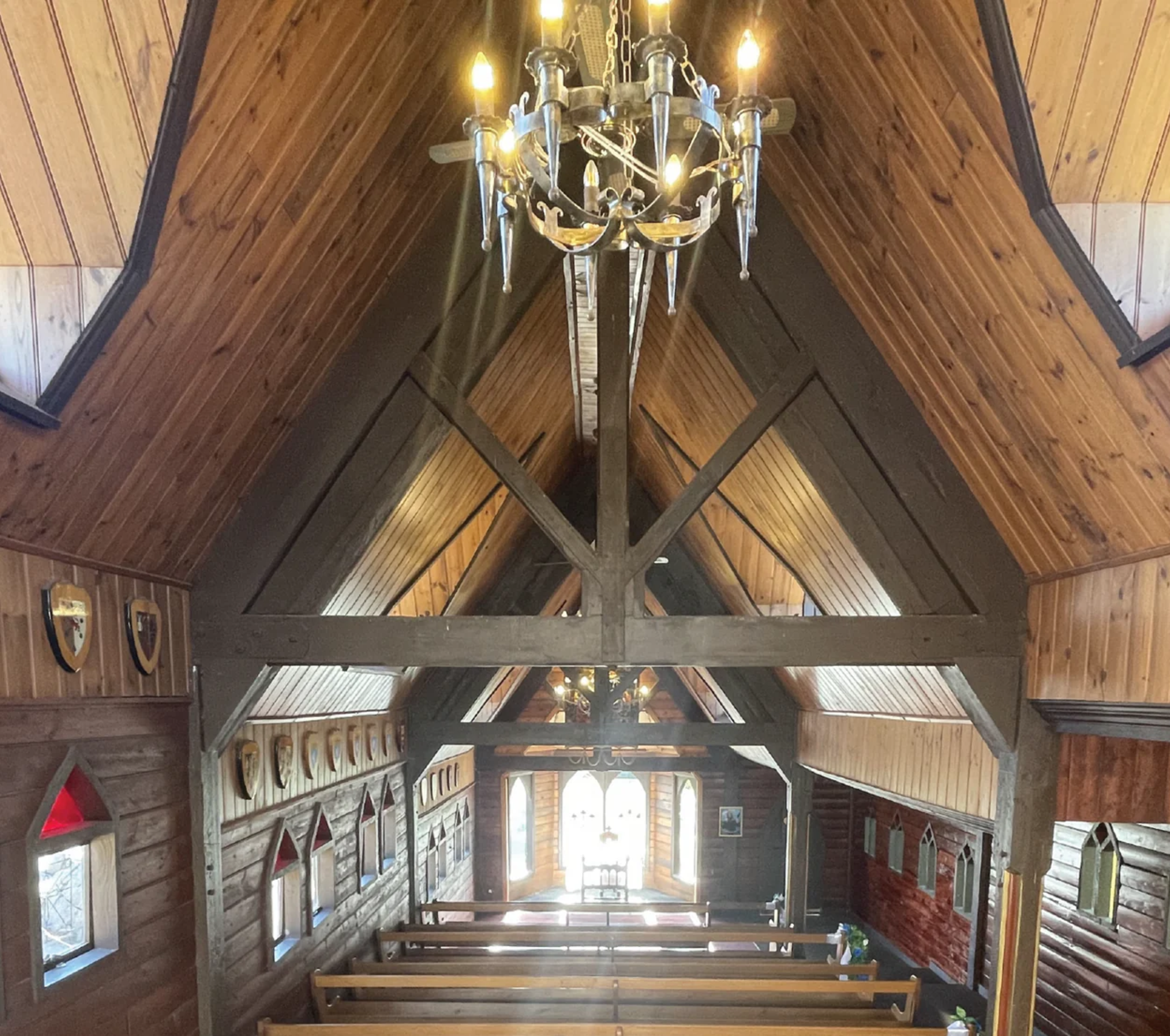 Interior of a rustic wooden chapel with chandeliers and pews facing bright arched windows.