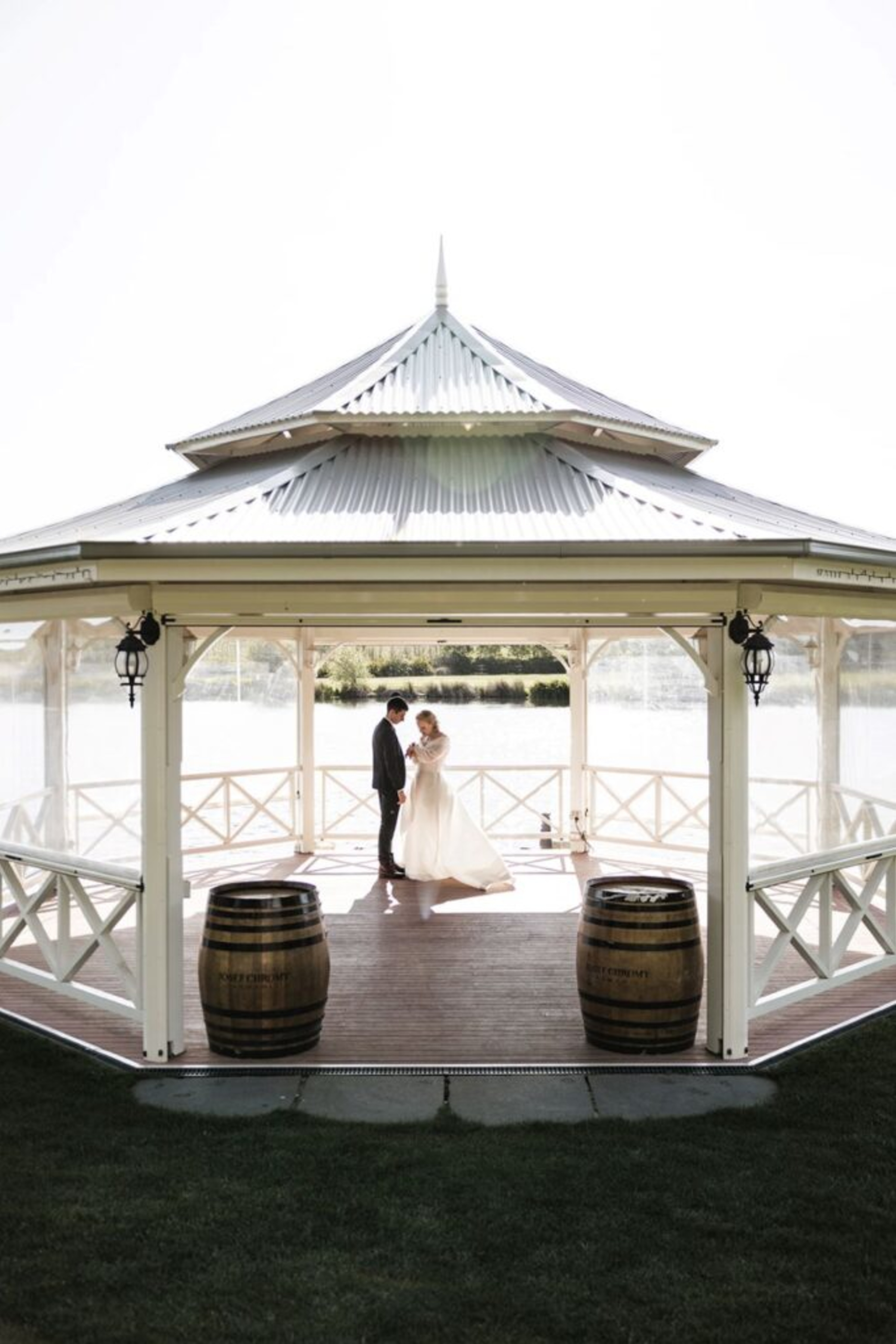 Bride and groom share a quiet moment inside a lakeside gazebo with wooden barrels and soft natural light.