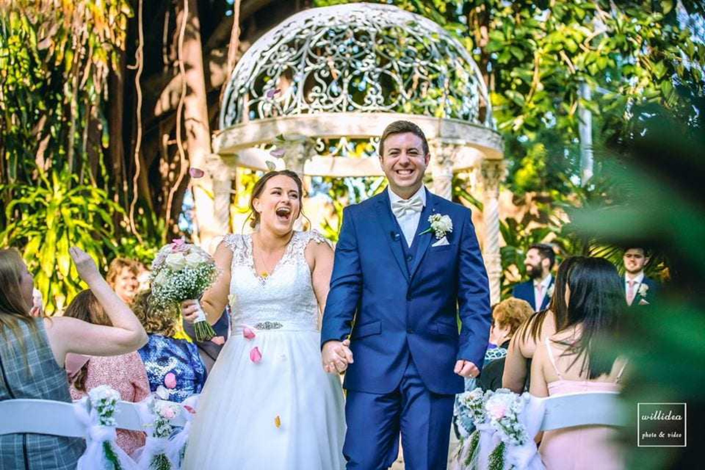 Joyful bride and groom walk down an outdoor garden aisle just married under a decorative gazebo.