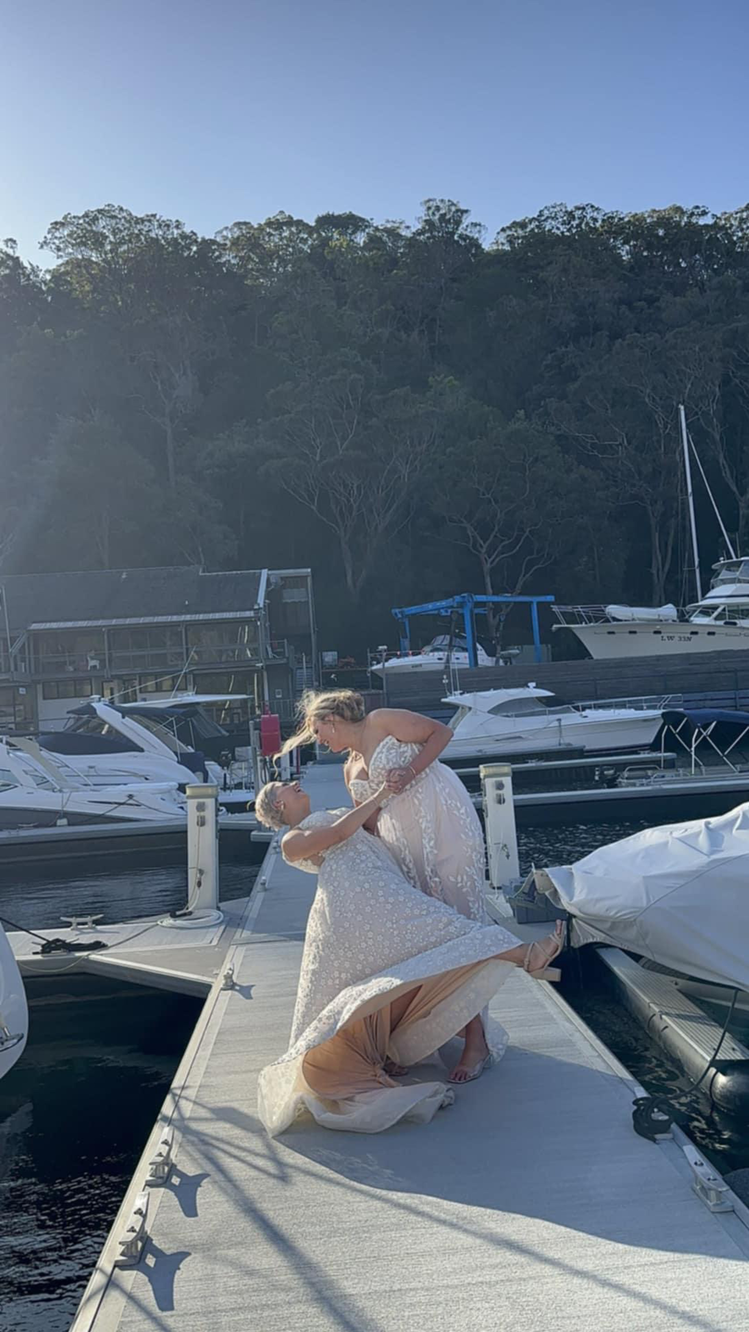 Two brides in wedding dresses share a romantic dip on a sunny marina dock surrounded by boats.