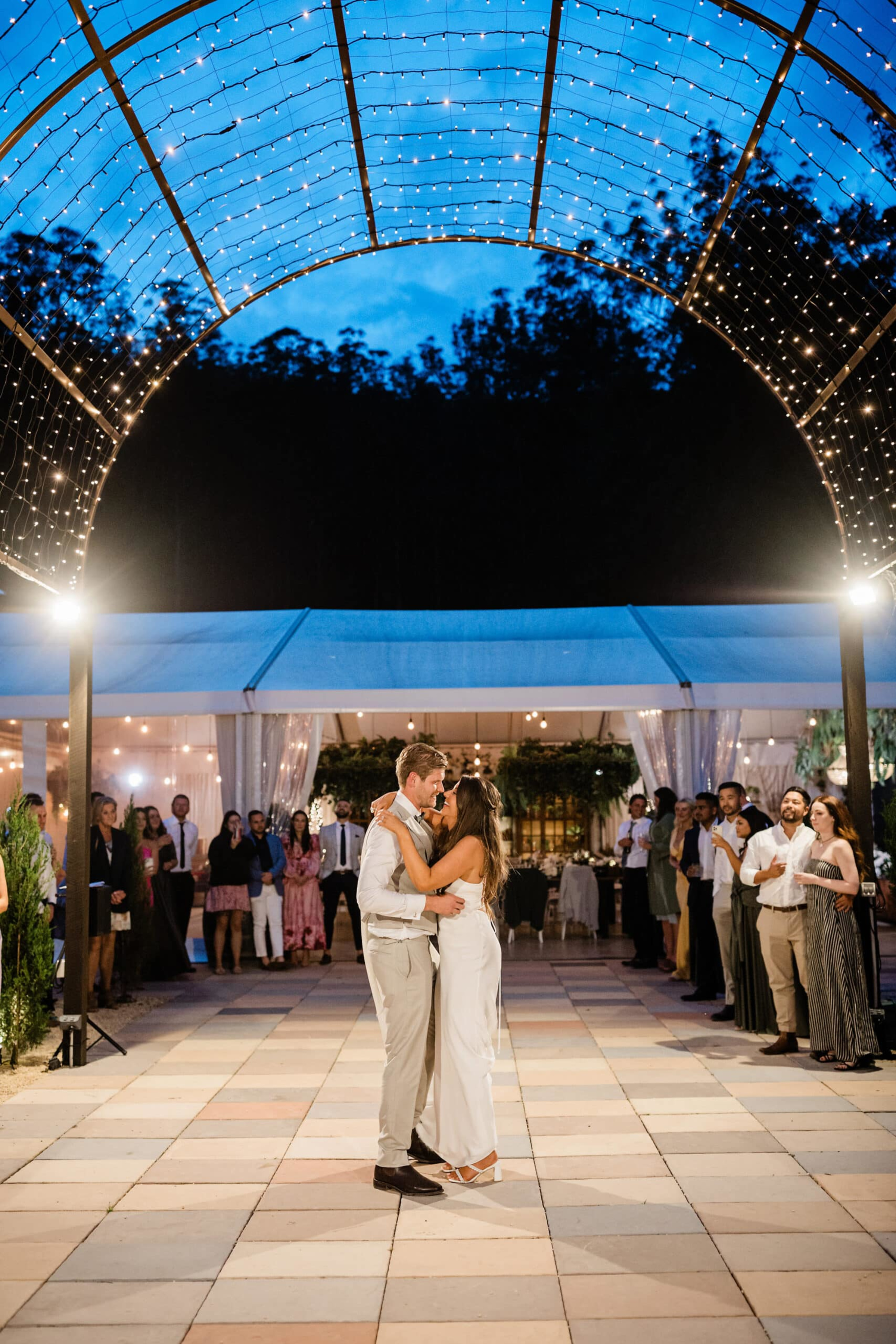 Couple shares their first dance under twinkling string lights at an outdoor evening wedding reception.