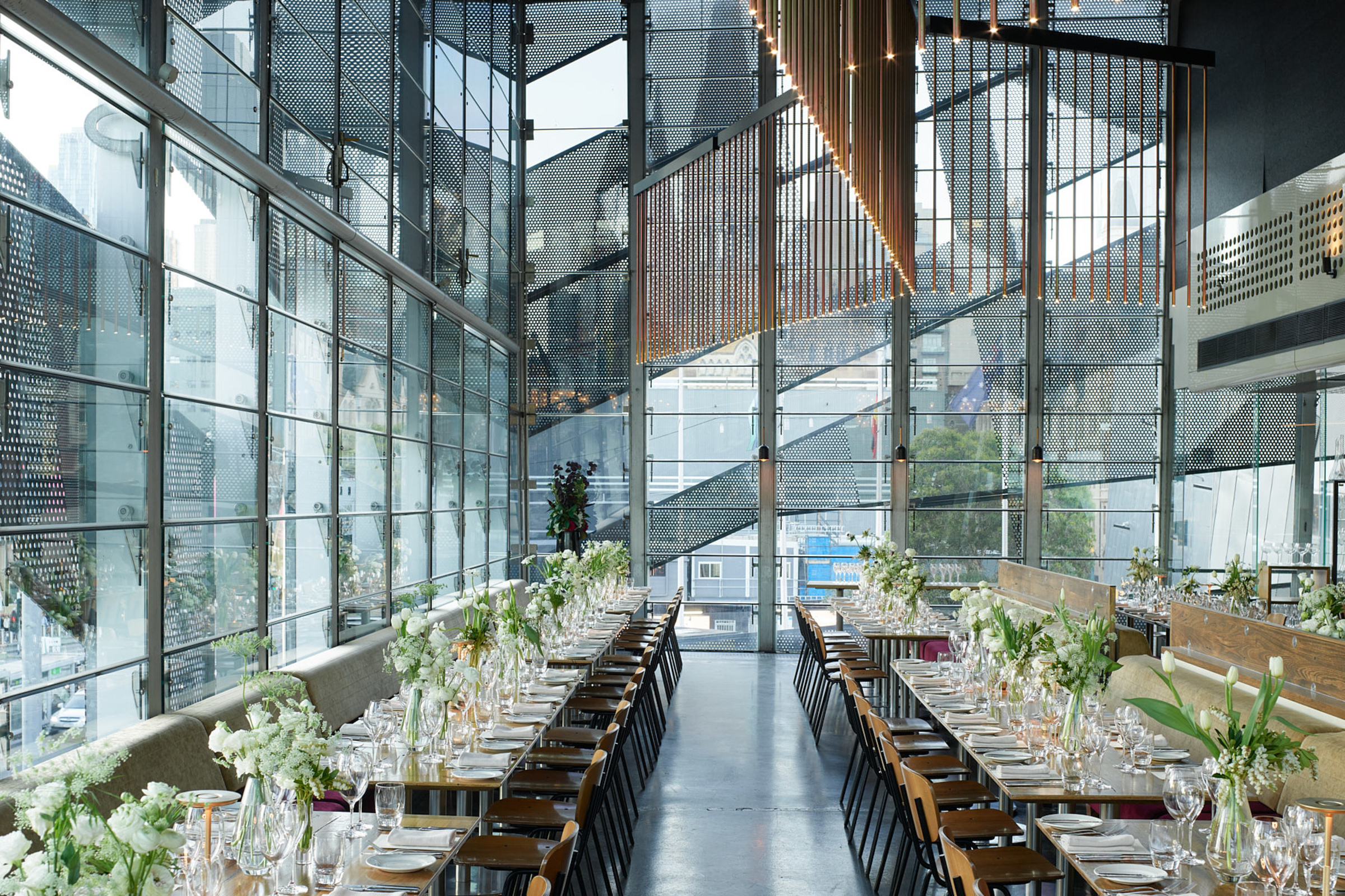 Modern glass-walled wedding reception venue with long banquet tables, white florals, and city views.