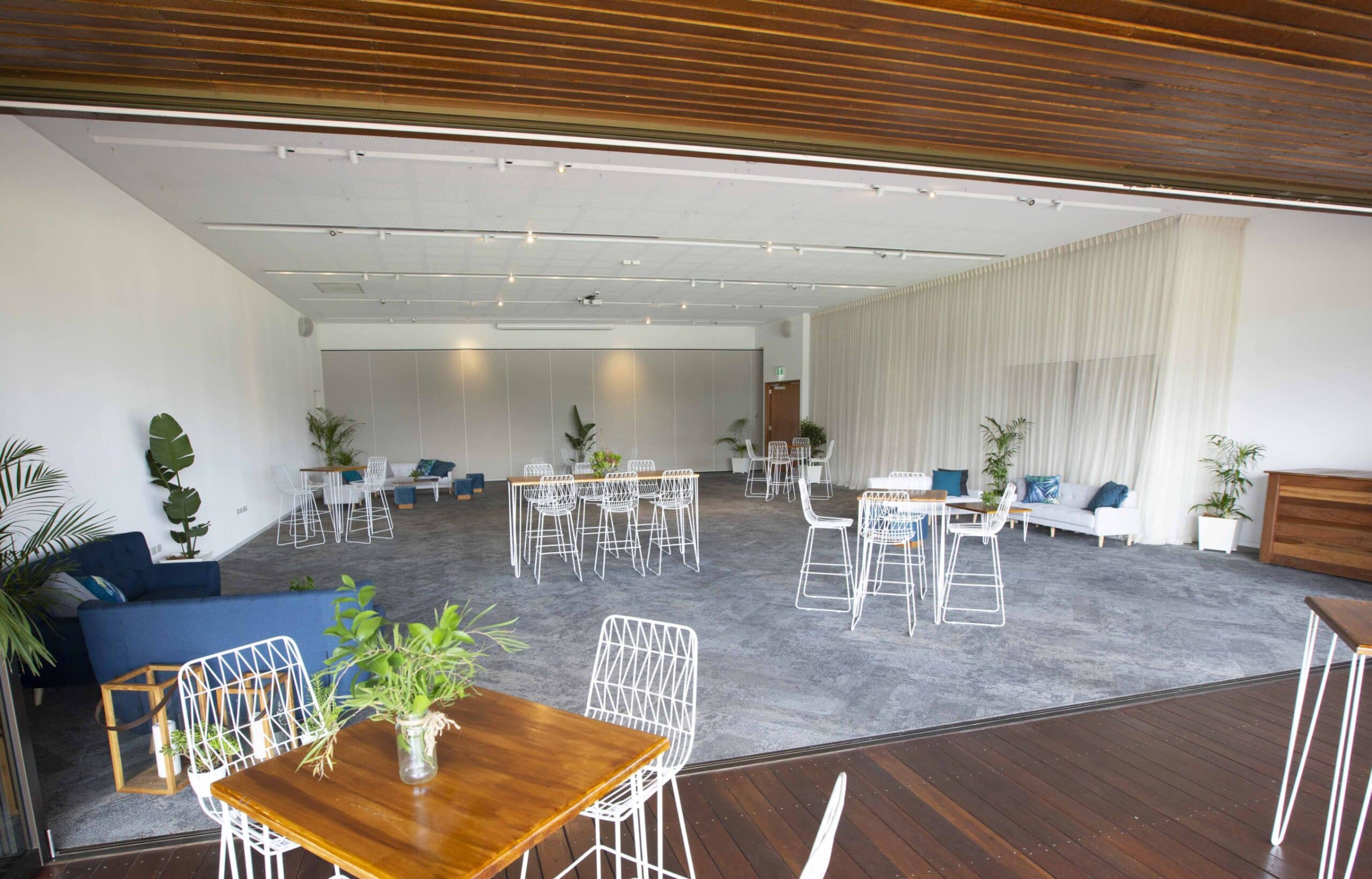 Spacious modern indoor wedding venue with white chairs, high tables, and lounge seating accented by greenery.