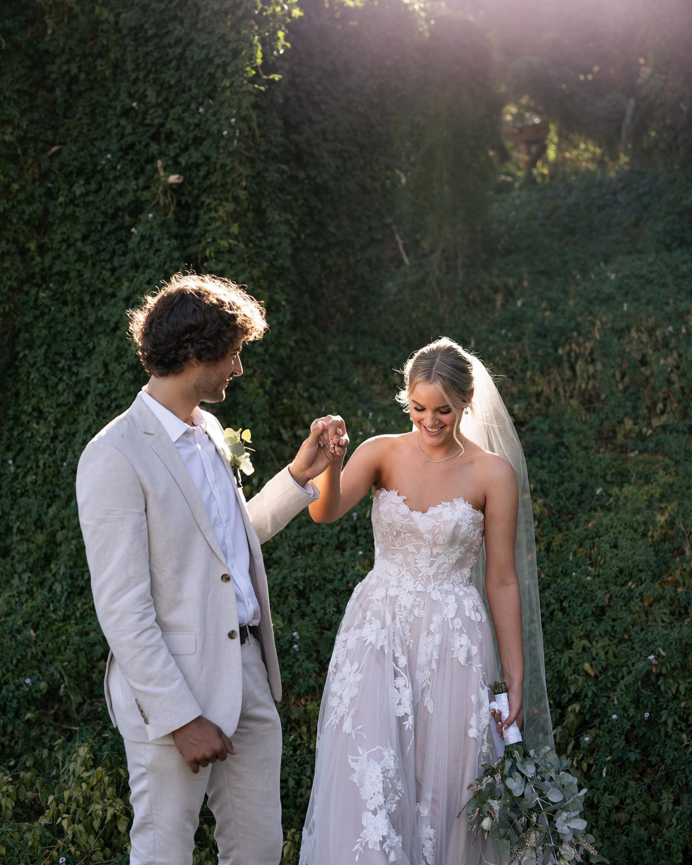 Bride and groom share a tender moment outdoors, holding hands in a sunlit, ivy-covered garden.
