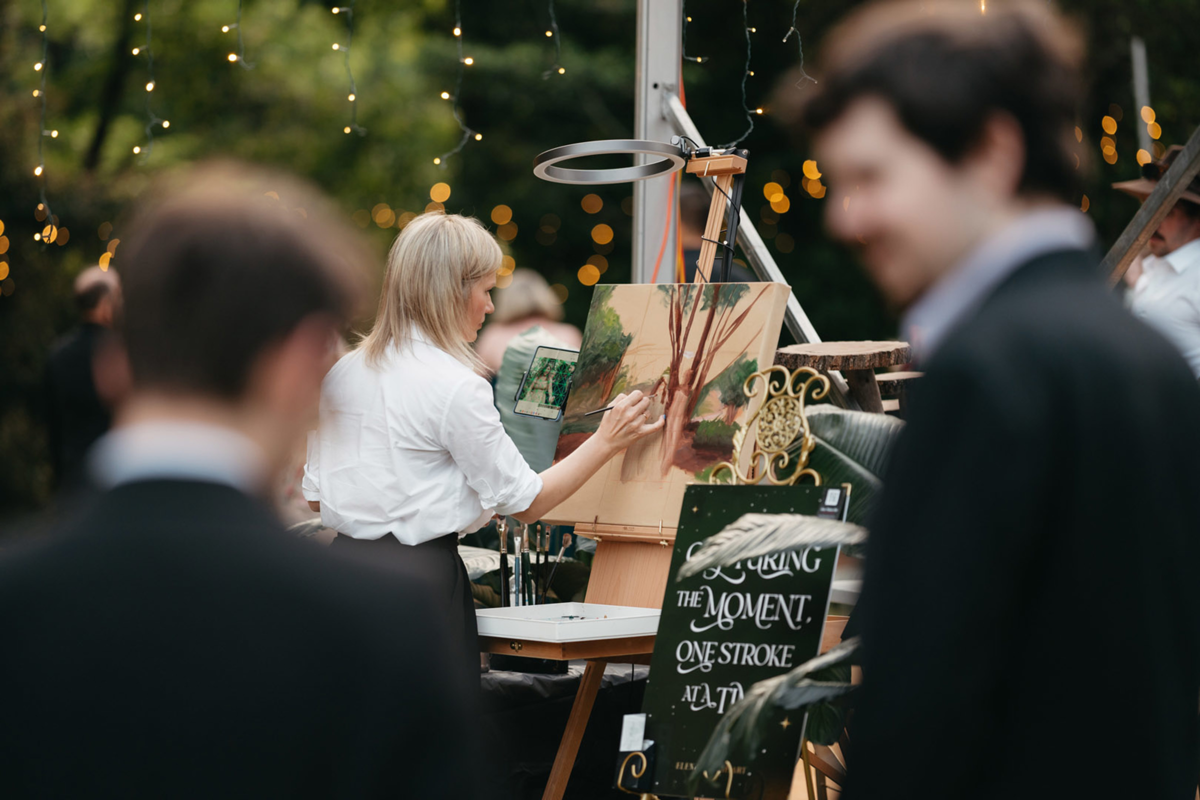Live painter creates an artwork at an outdoor wedding reception under string lights with guests nearby.