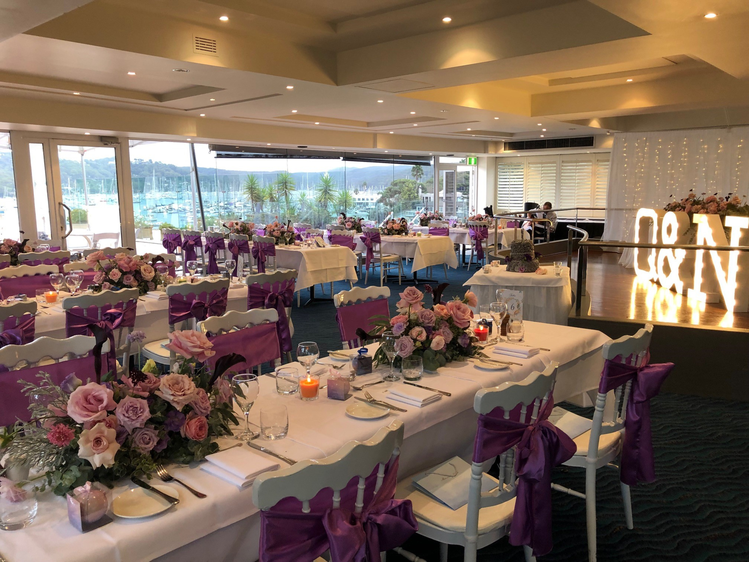 Waterfront wedding reception with purple floral centerpieces, chair sashes, and glowing marquee initials.