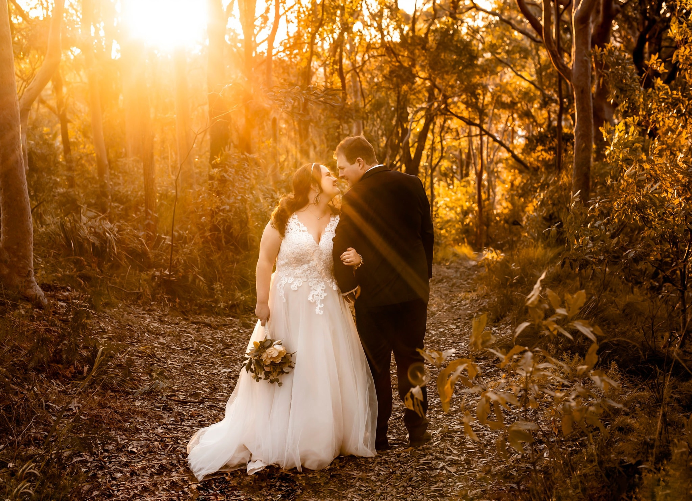 Bride and groom share a kiss on a sunlit forest path during golden hour.