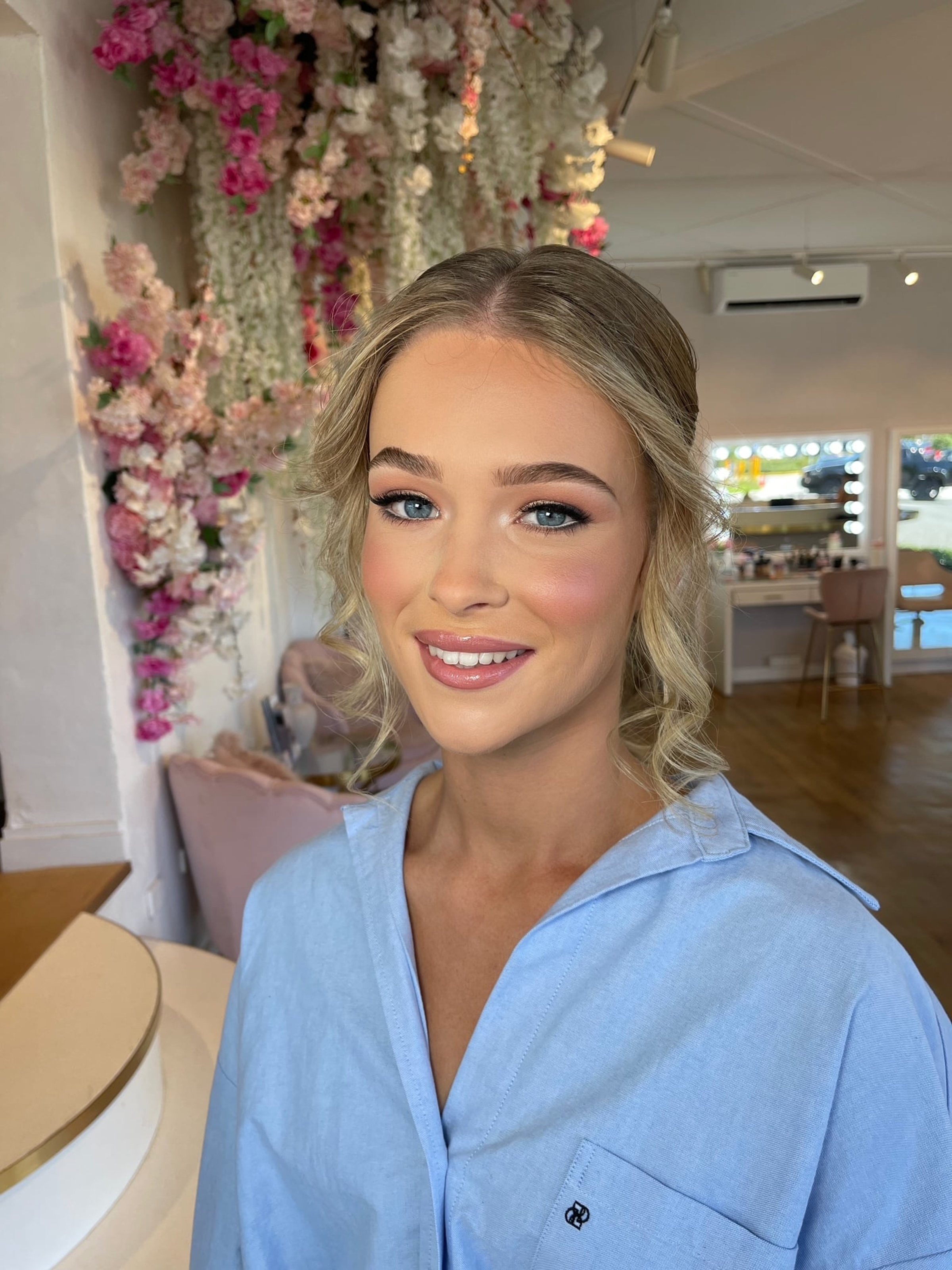 Smiling woman with soft glam bridal makeup and loose updo in a studio with pink floral backdrop.