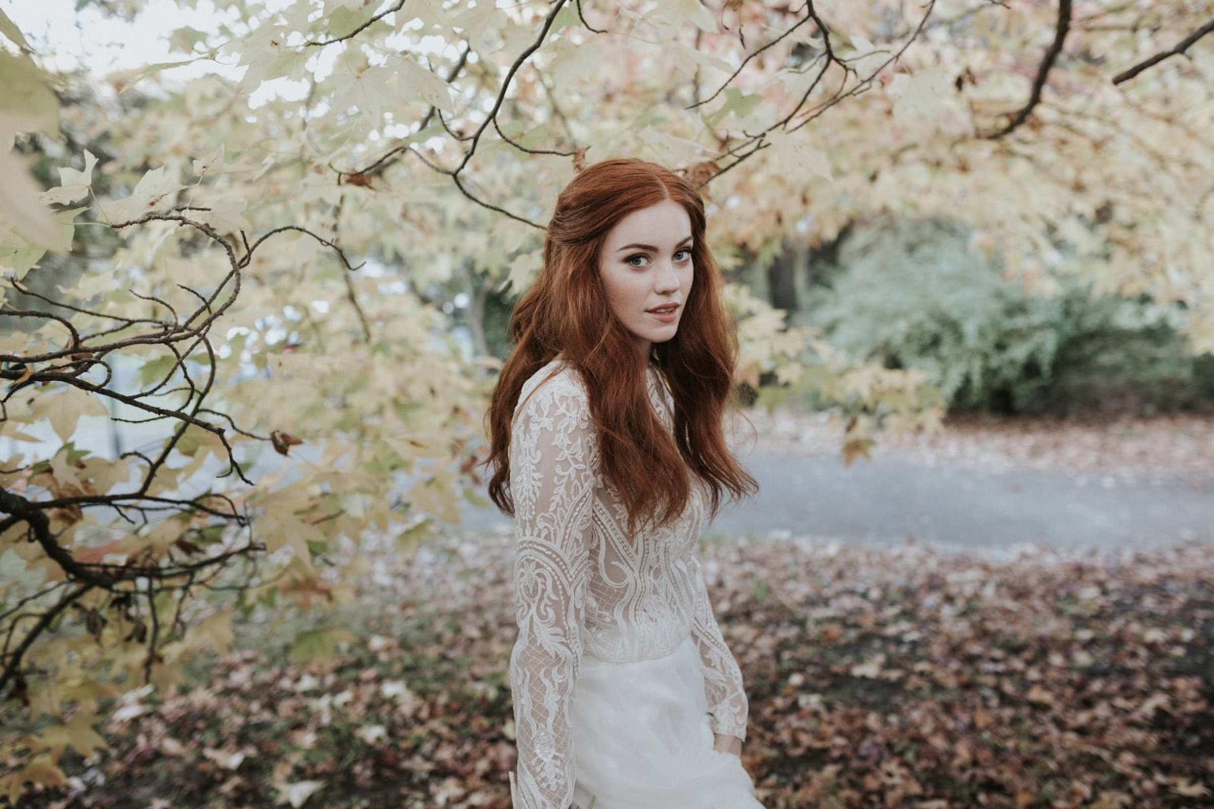 Bride in a lace wedding dress poses outdoors among autumn leaves and tree branches.