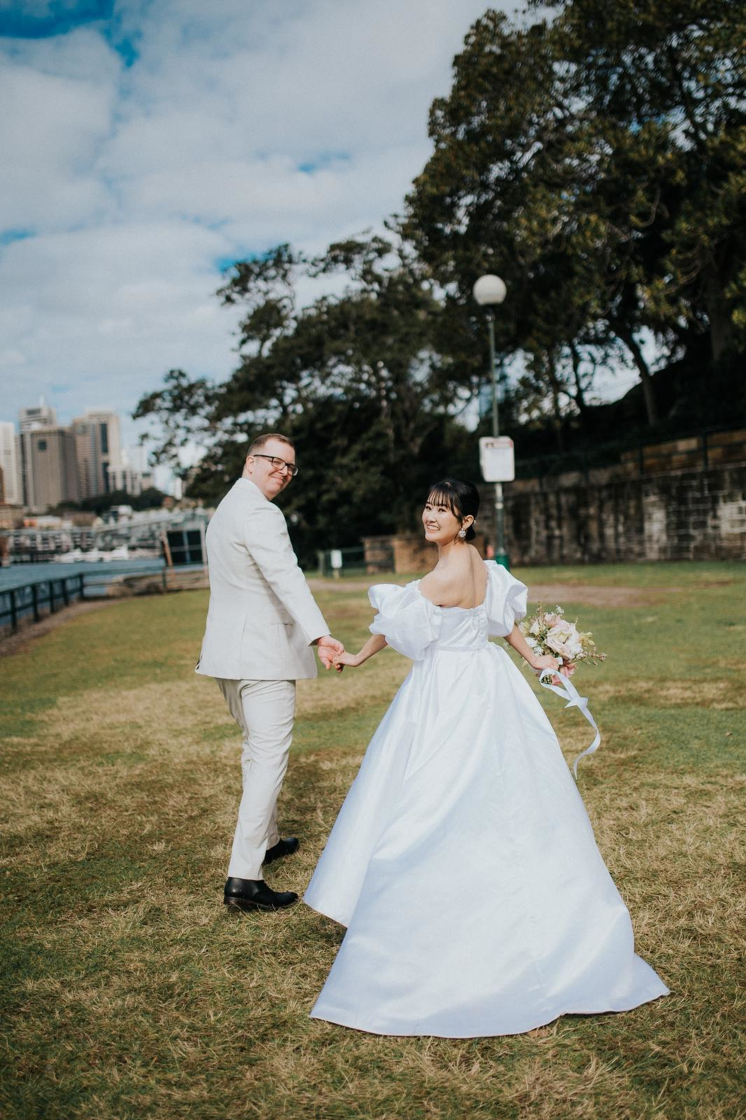 Newlywed couple holding hands and walking through a grassy park with city skyline in the background.