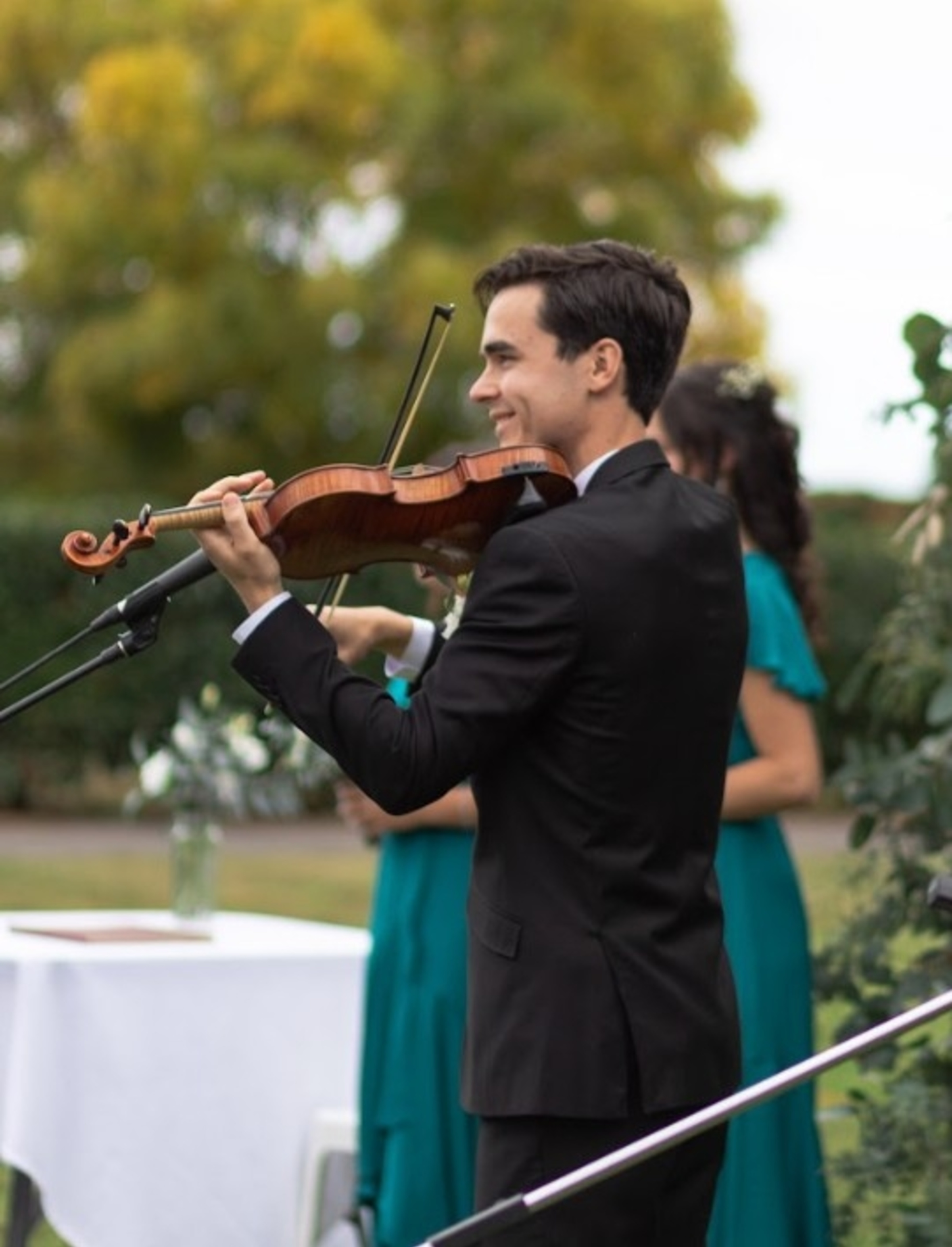 Smiling violinist in a black suit plays live music at an outdoor wedding ceremony.