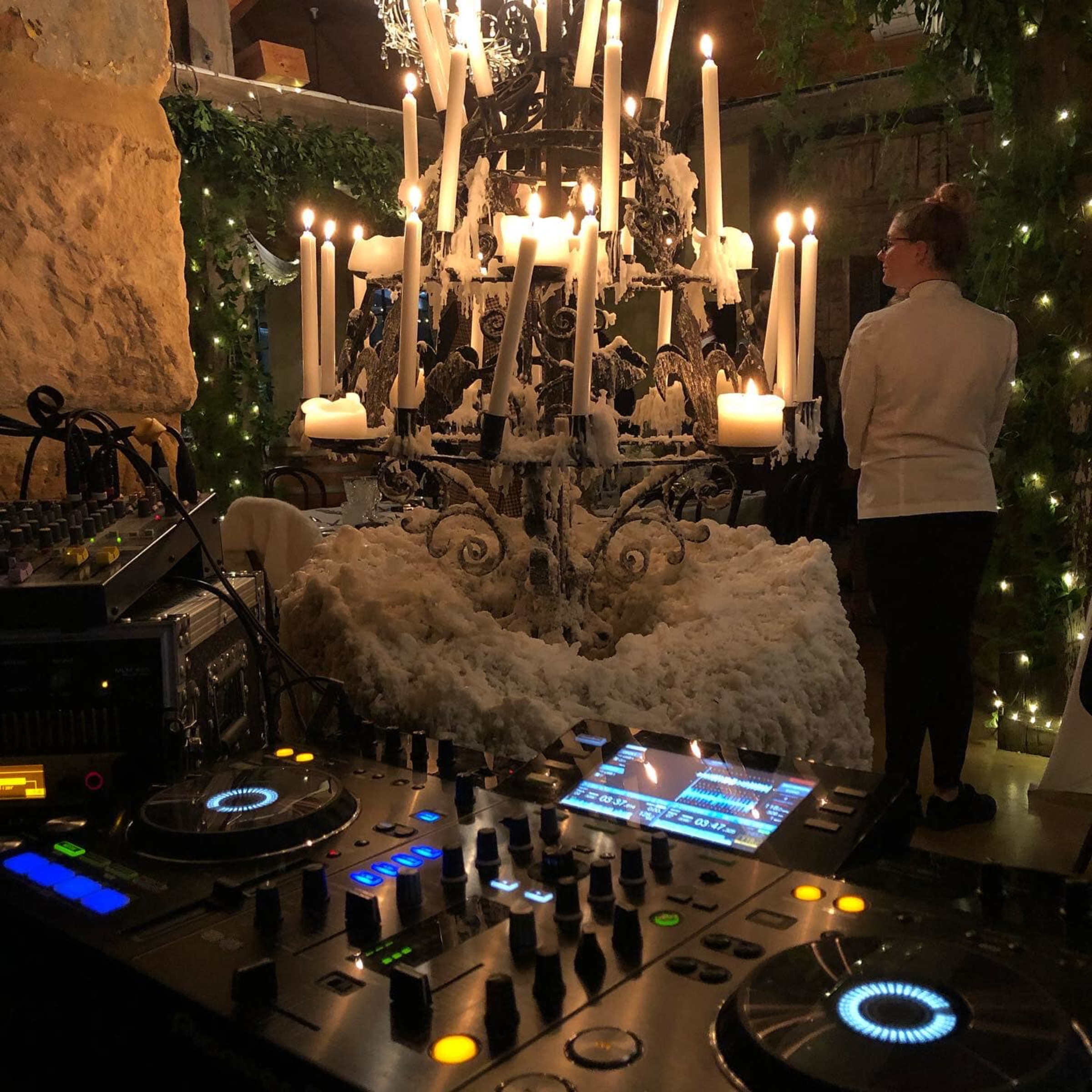 DJ booth set up beside a large candlelit chandelier with snowy decor and twinkling lights at an indoor wedding reception.