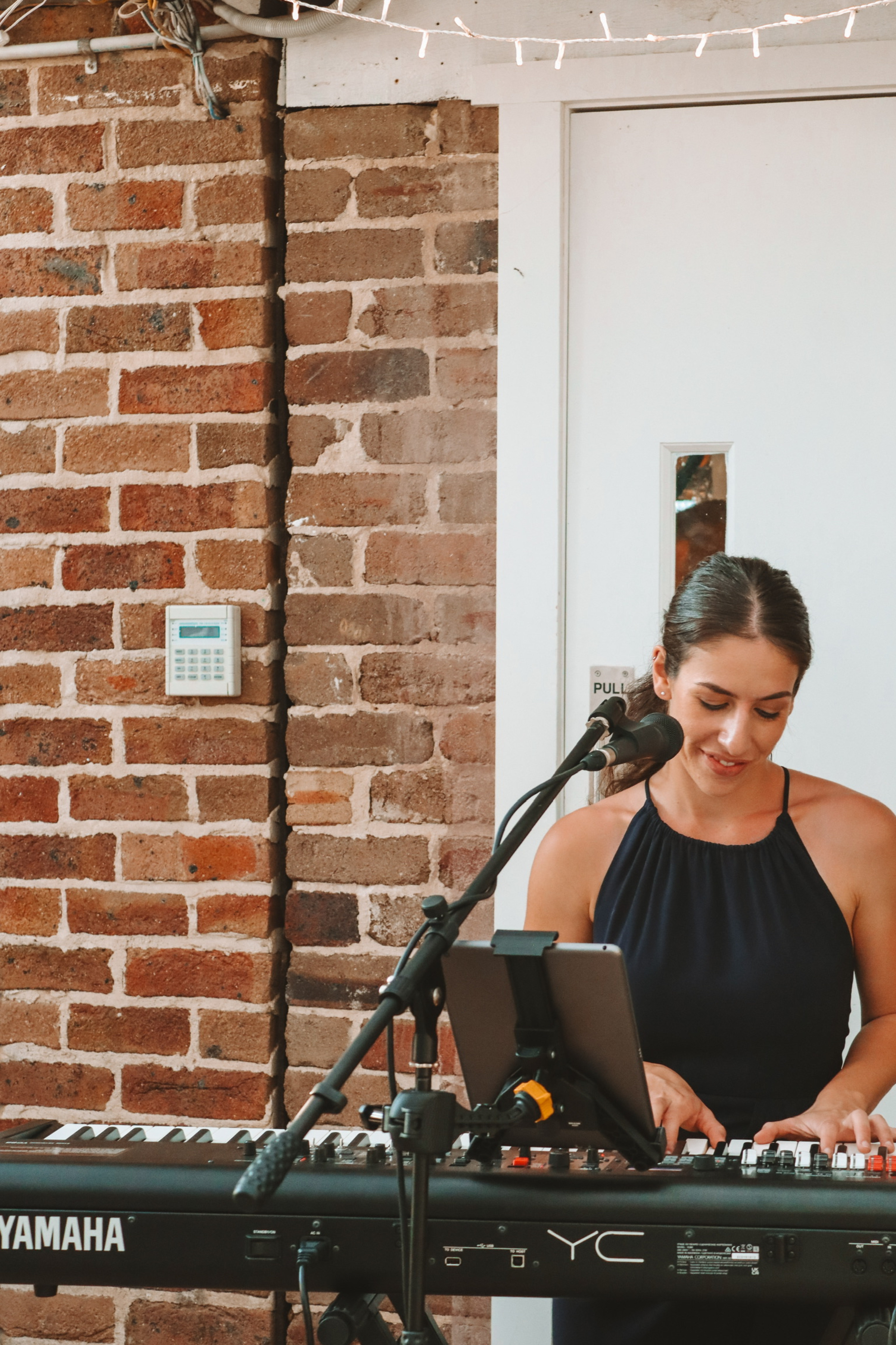 Female musician plays keyboard and sings into a microphone against a brick wall at a wedding event.