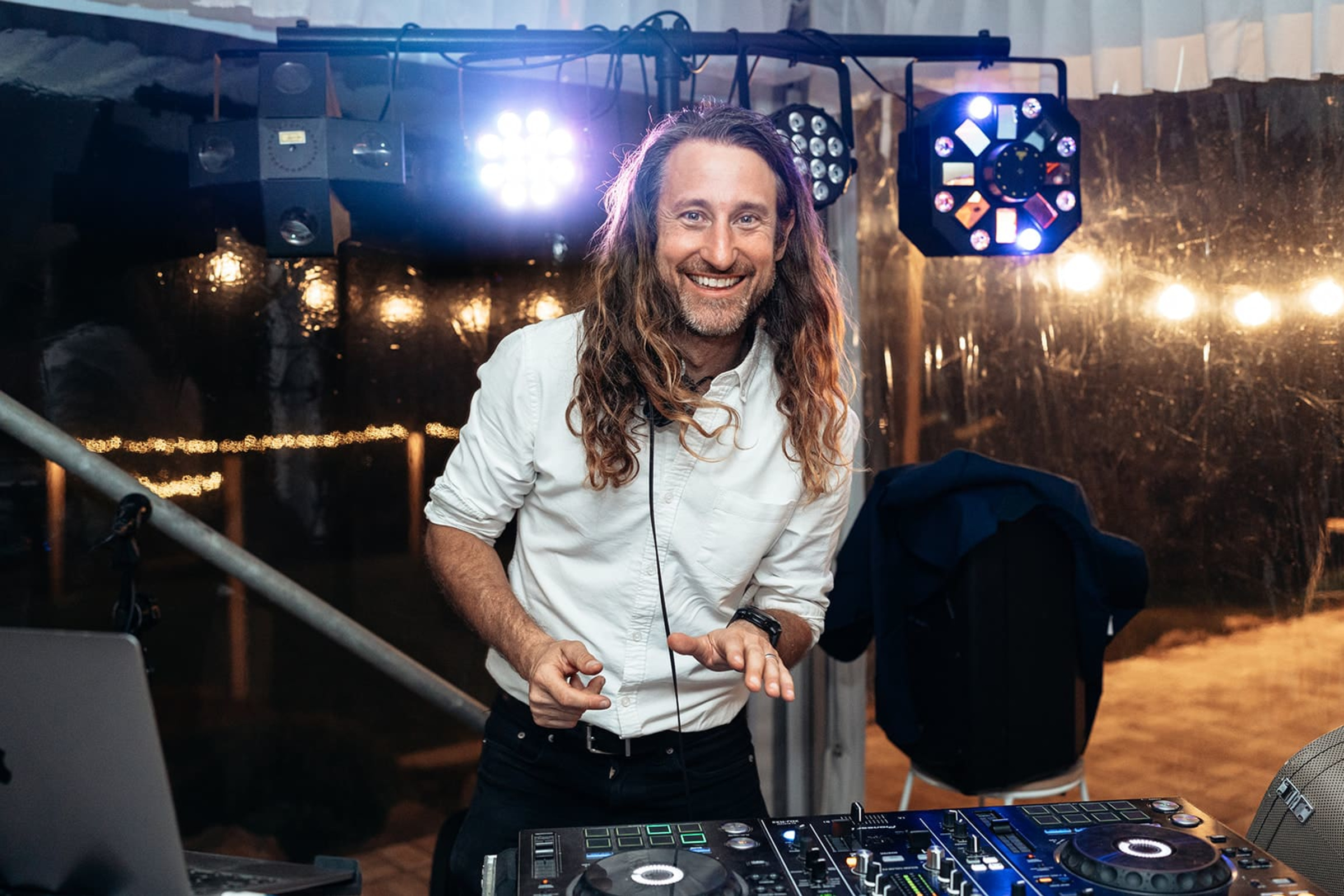Smiling wedding DJ mixing music behind a lit DJ booth at a nighttime reception.