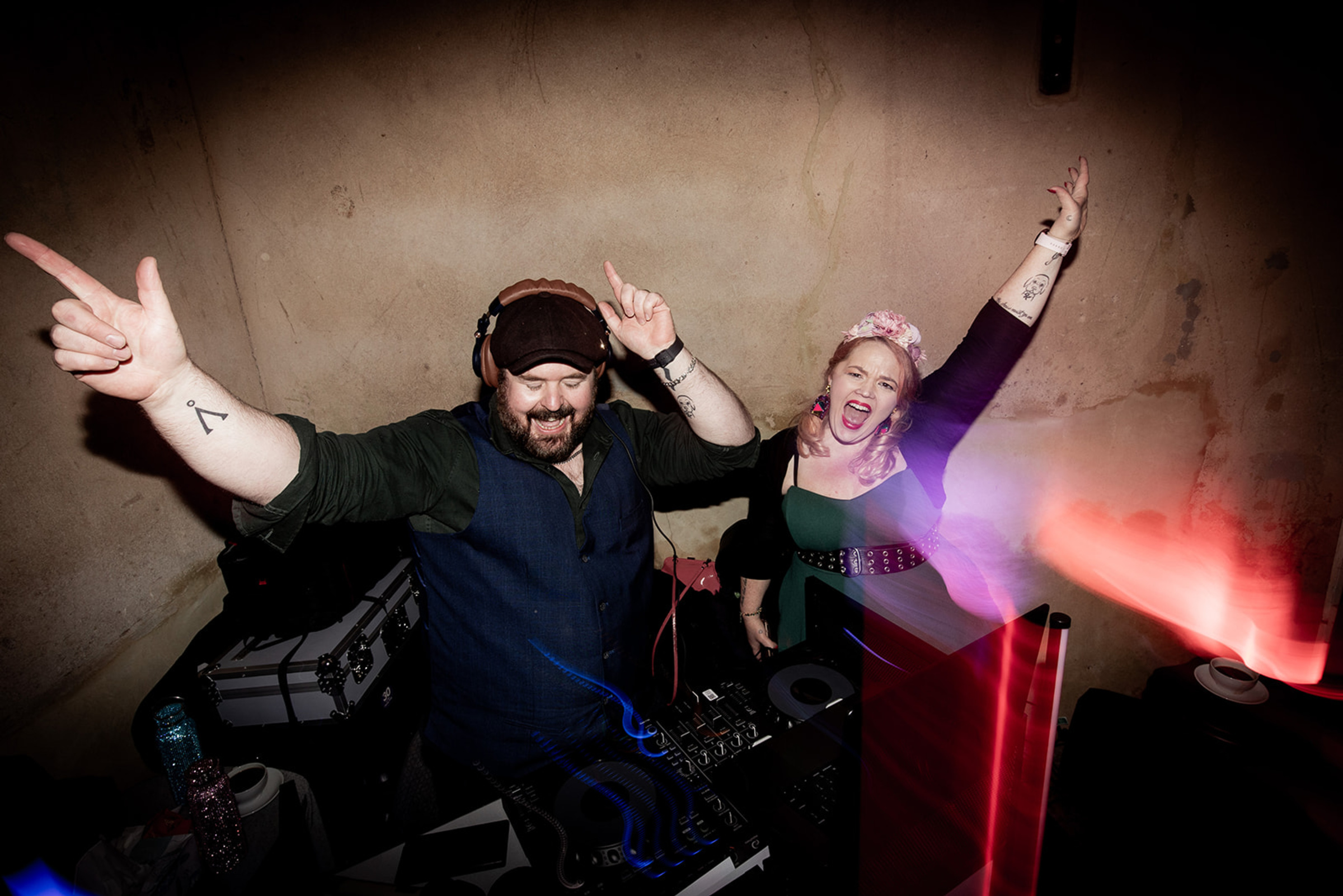 Two energetic DJs celebrate behind the booth with colorful party lights at a wedding reception.