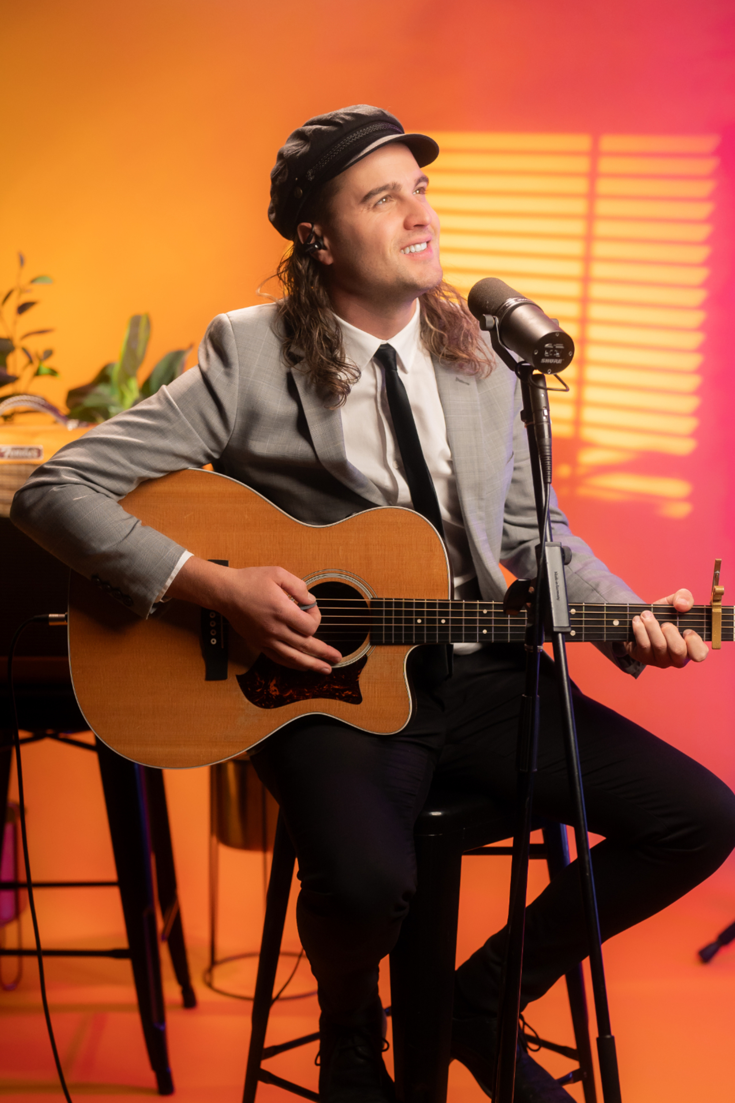 Smiling guitarist in a suit performs into a microphone against a warm orange and pink backdrop.