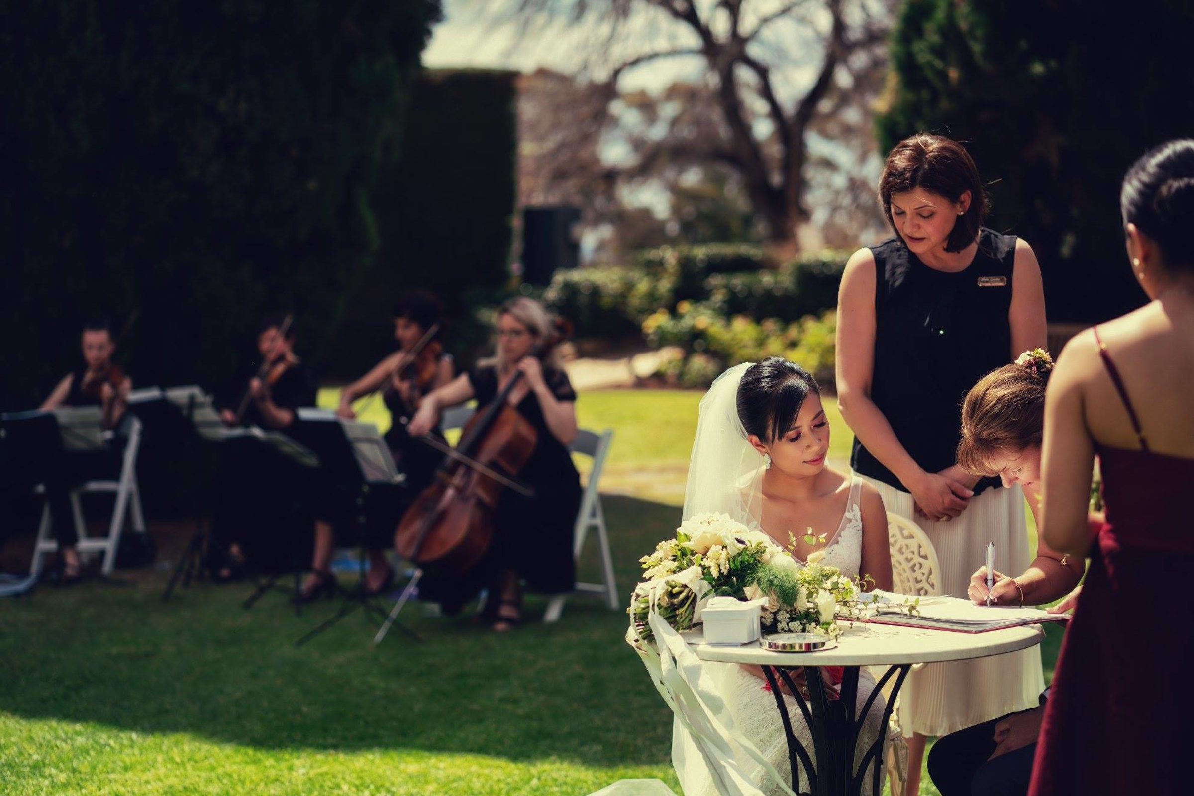 Bride signs the marriage certificate at an outdoor garden ceremony with a live string quartet in the background.