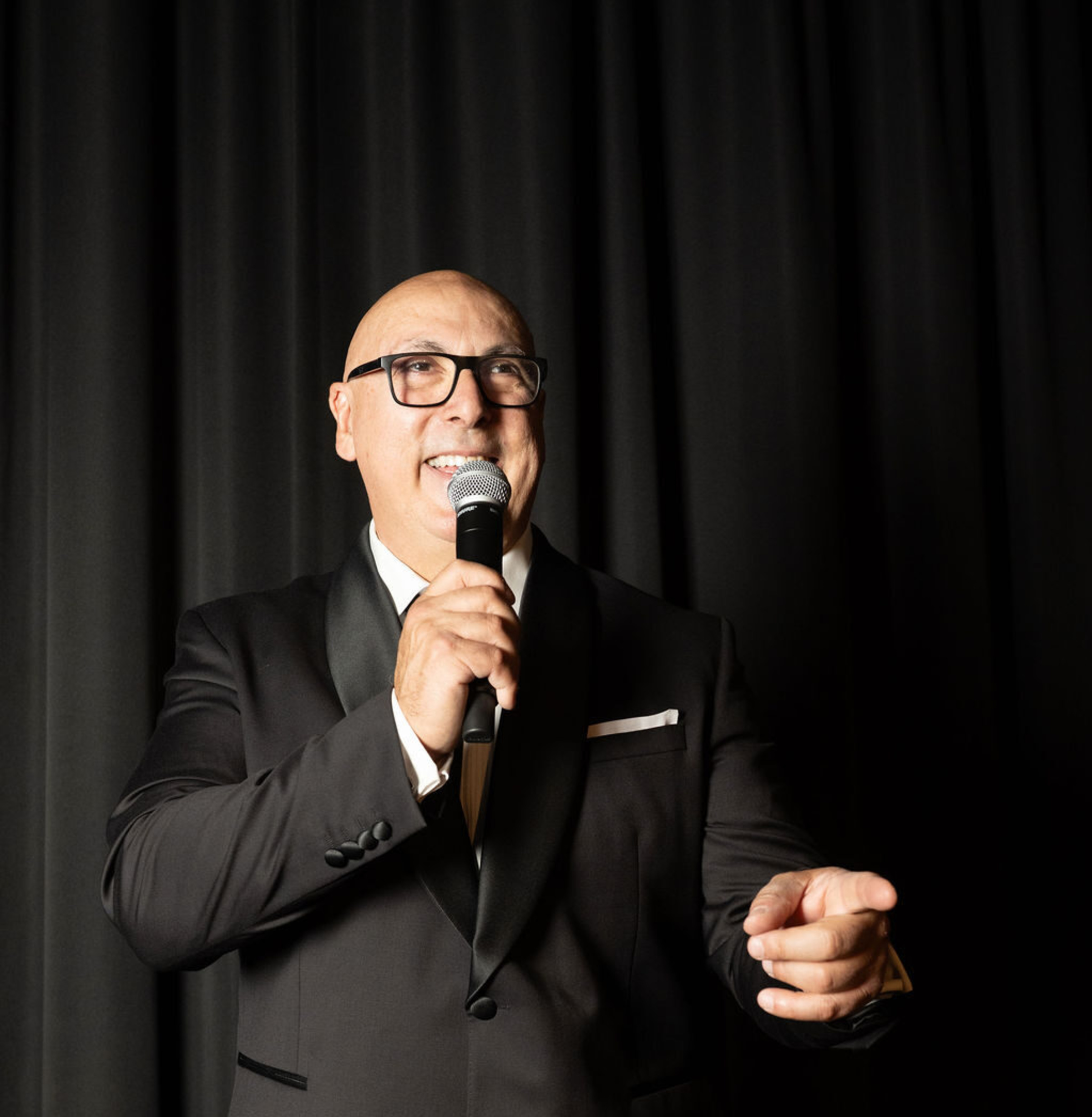 Smiling wedding emcee in a black tuxedo speaking into a microphone against a dark curtain backdrop.