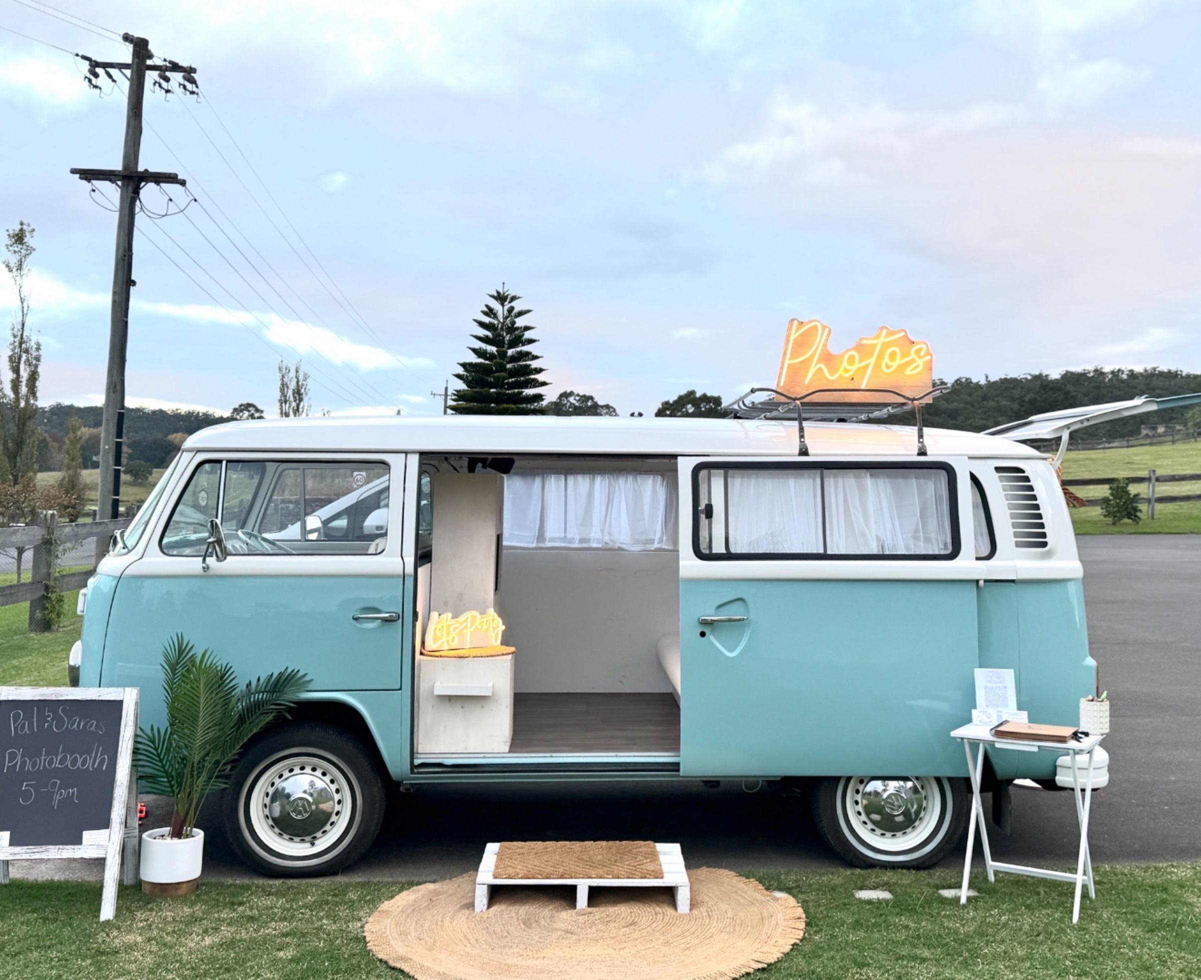 Vintage teal camper van photo booth setup at an outdoor wedding venue with neon sign and decor.