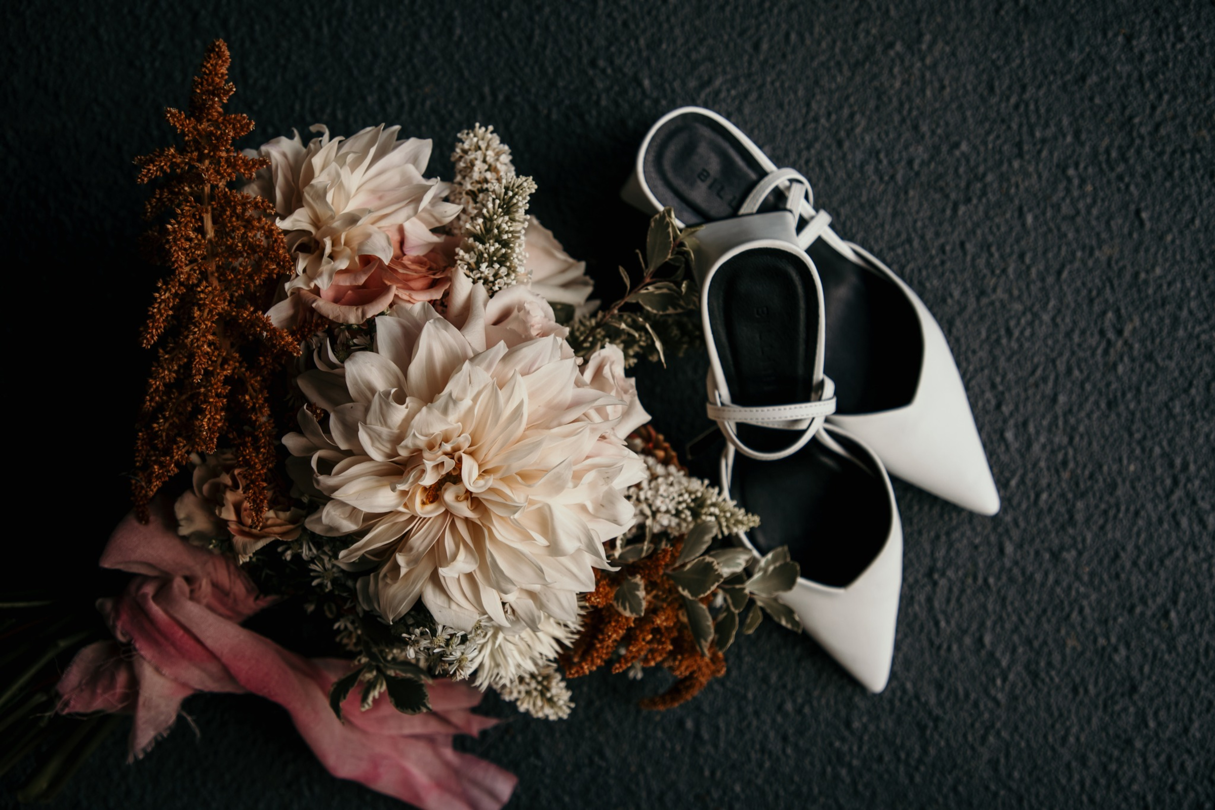 Flatlay of white bridal shoes beside a romantic bouquet of neutral and blush flowers on a dark surface.