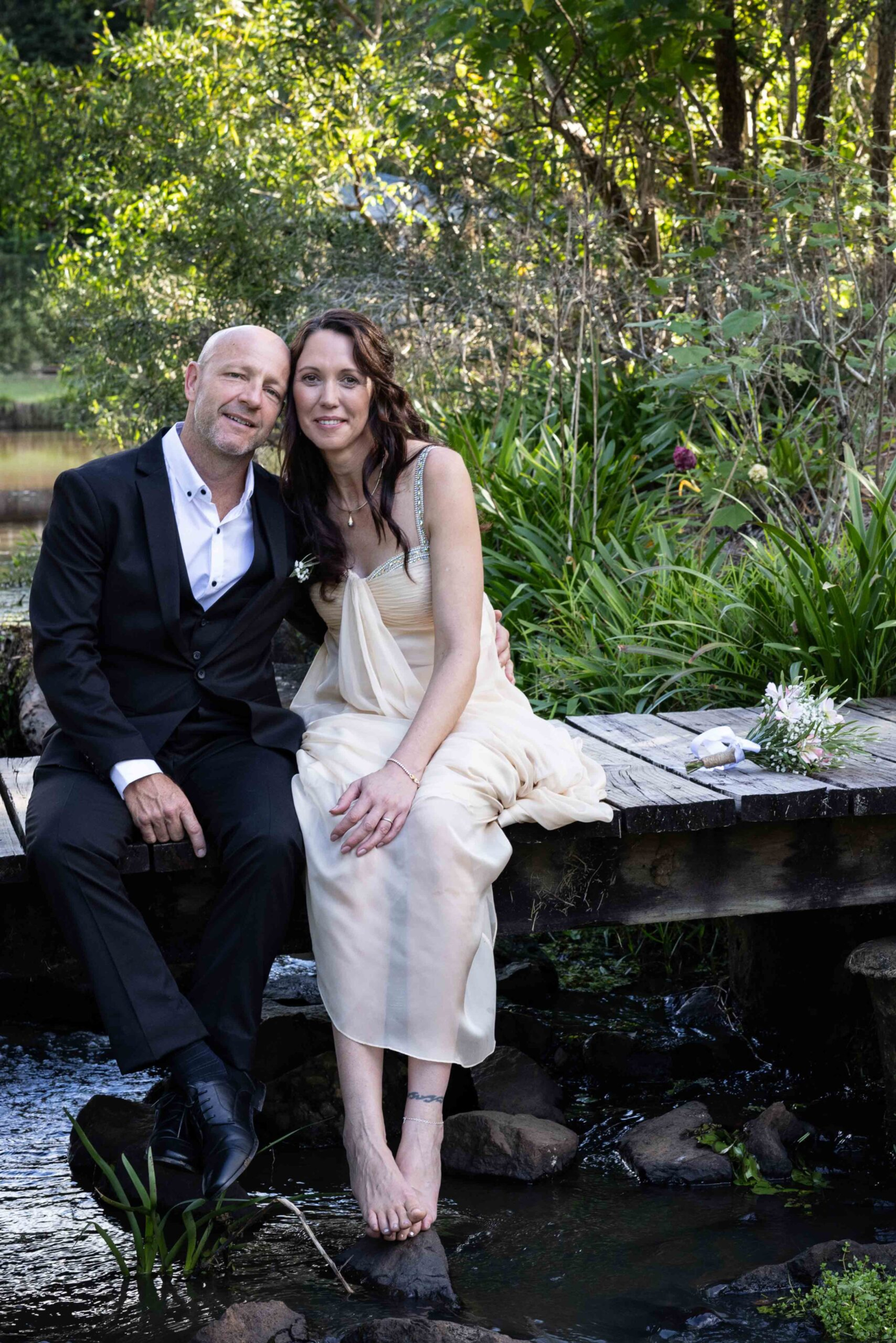 Bride and groom sit barefoot on a wooden bridge over a stream in a lush green garden.