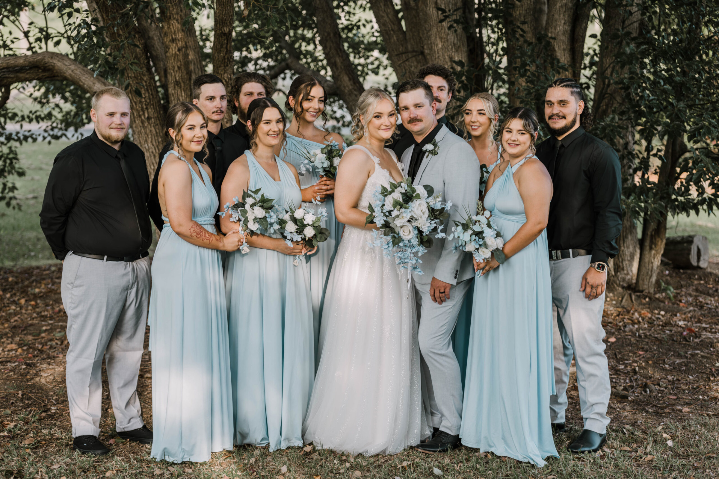 Bride and groom posing outdoors with bridesmaids in light blue dresses and groomsmen under large trees.