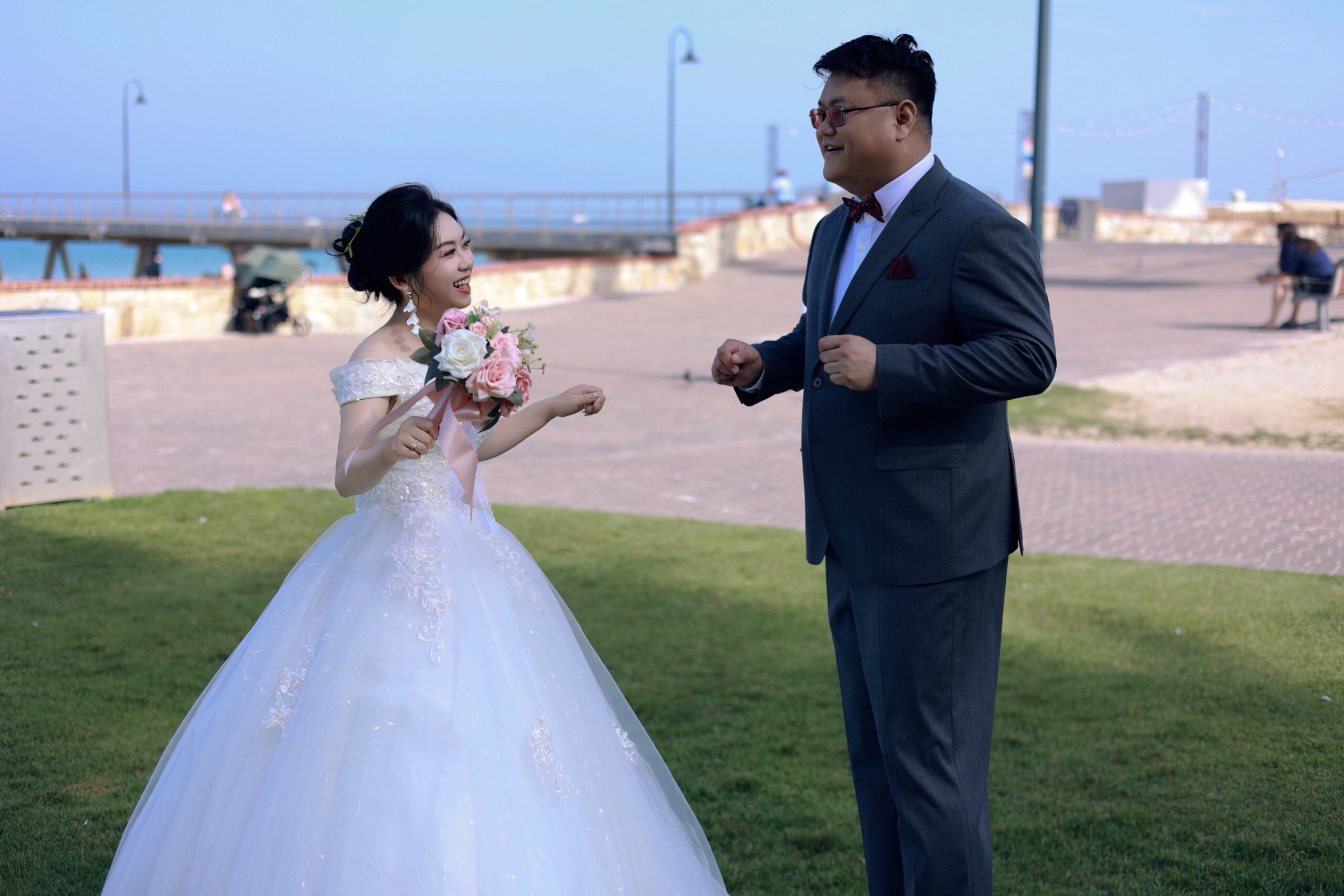 Bride and groom share a joyful moment at a seaside lawn with bouquet in hand.