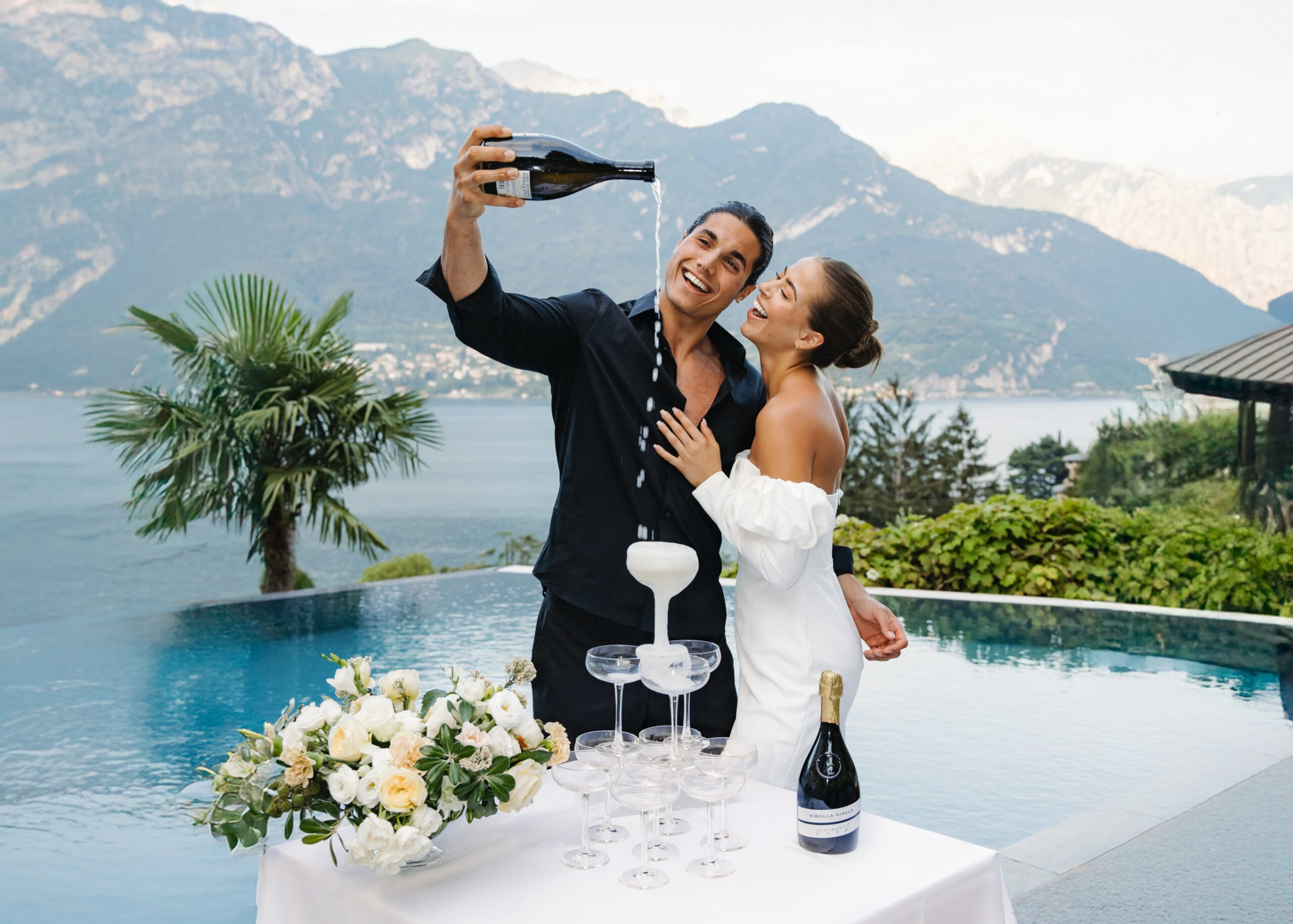 Joyful couple pours champagne into a tower by an infinity pool overlooking a scenic lake and mountains.