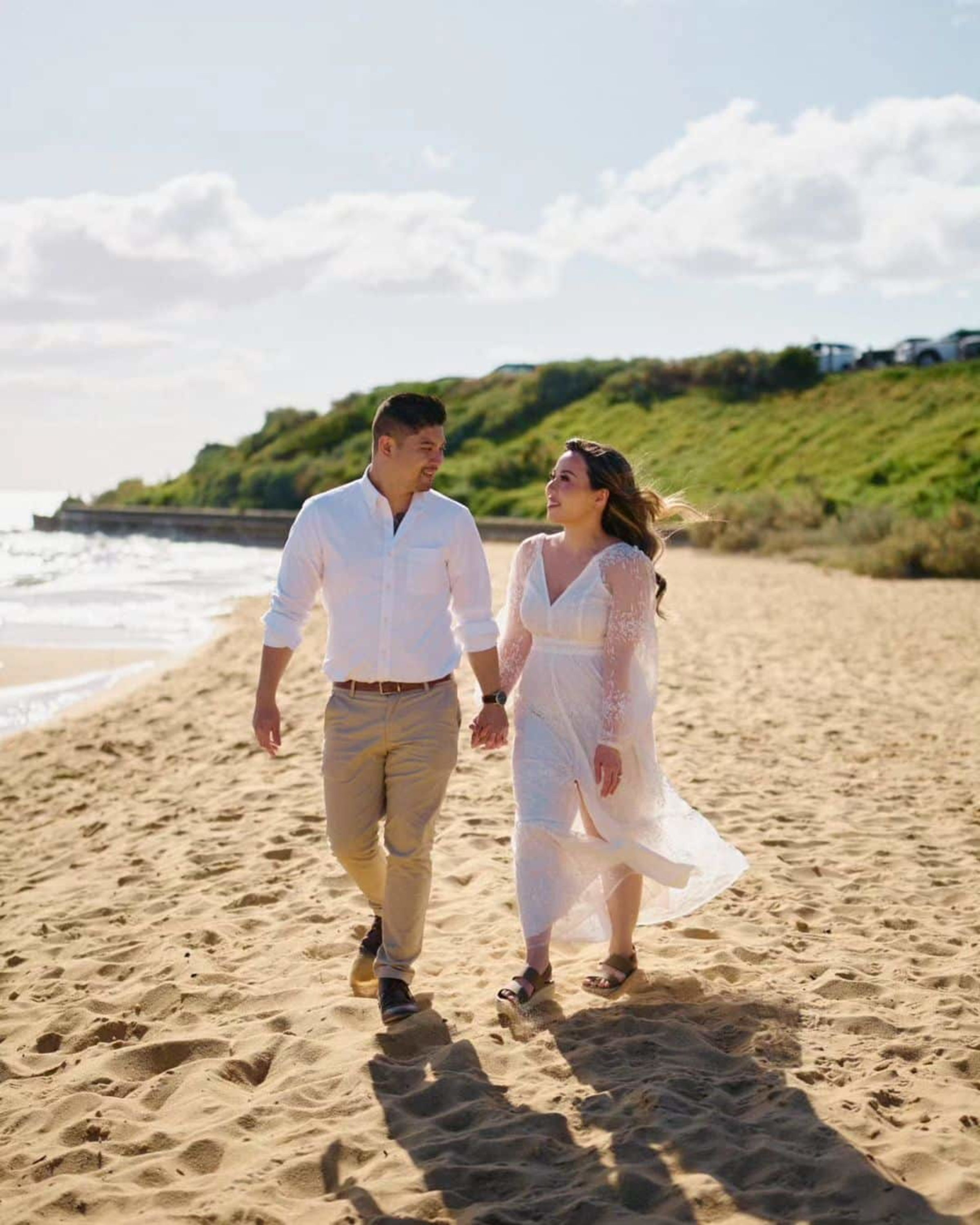A couple walks hand in hand along a sunny beach, dressed in light neutral outfits with waves and cliffs in the background.
