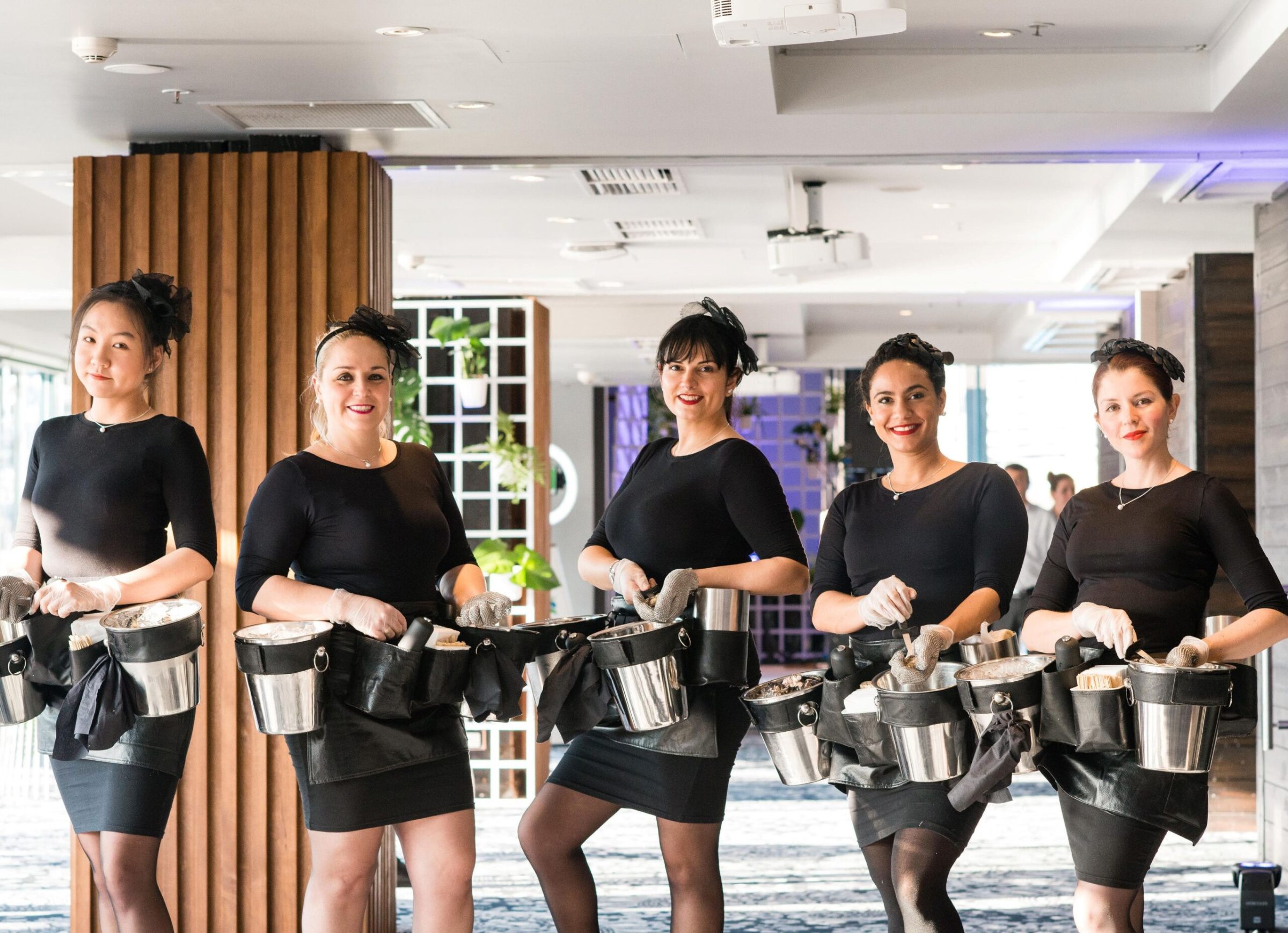 A line of smiling female catering staff in black outfits with service belts stands ready inside a bright wedding venue.
