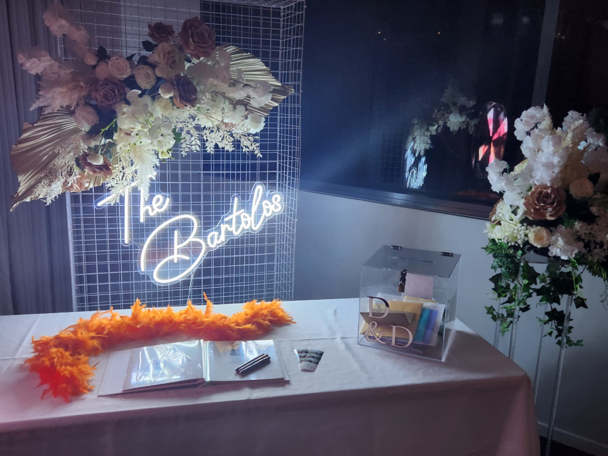 Wedding reception welcome table with neon surname sign, floral arrangements, guest book, and clear card box.