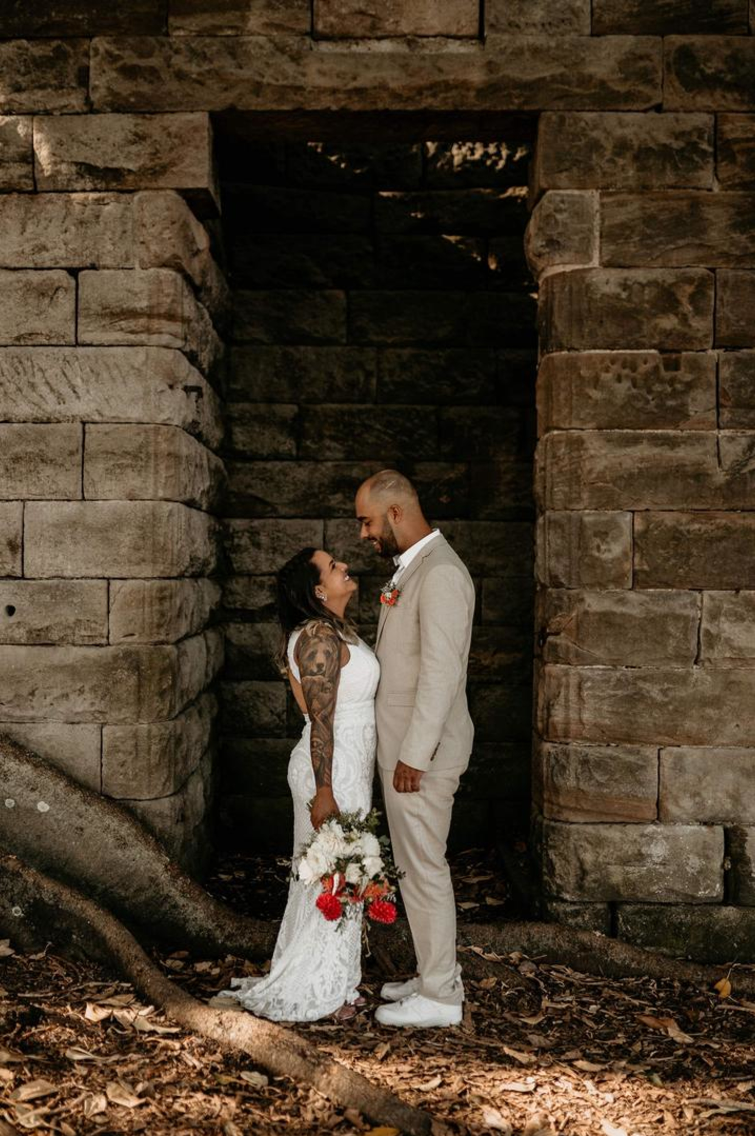 Bride and groom stand closely together under a rustic stone archway in a forest setting.