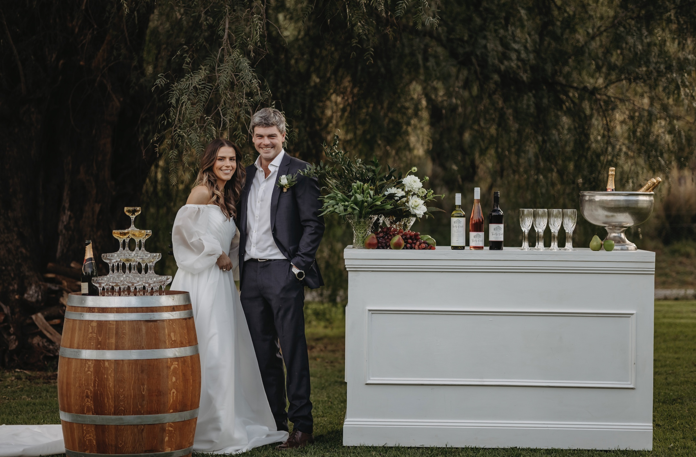 Bride and groom standing by an outdoor wedding bar with a champagne tower and wine display on a lawn.