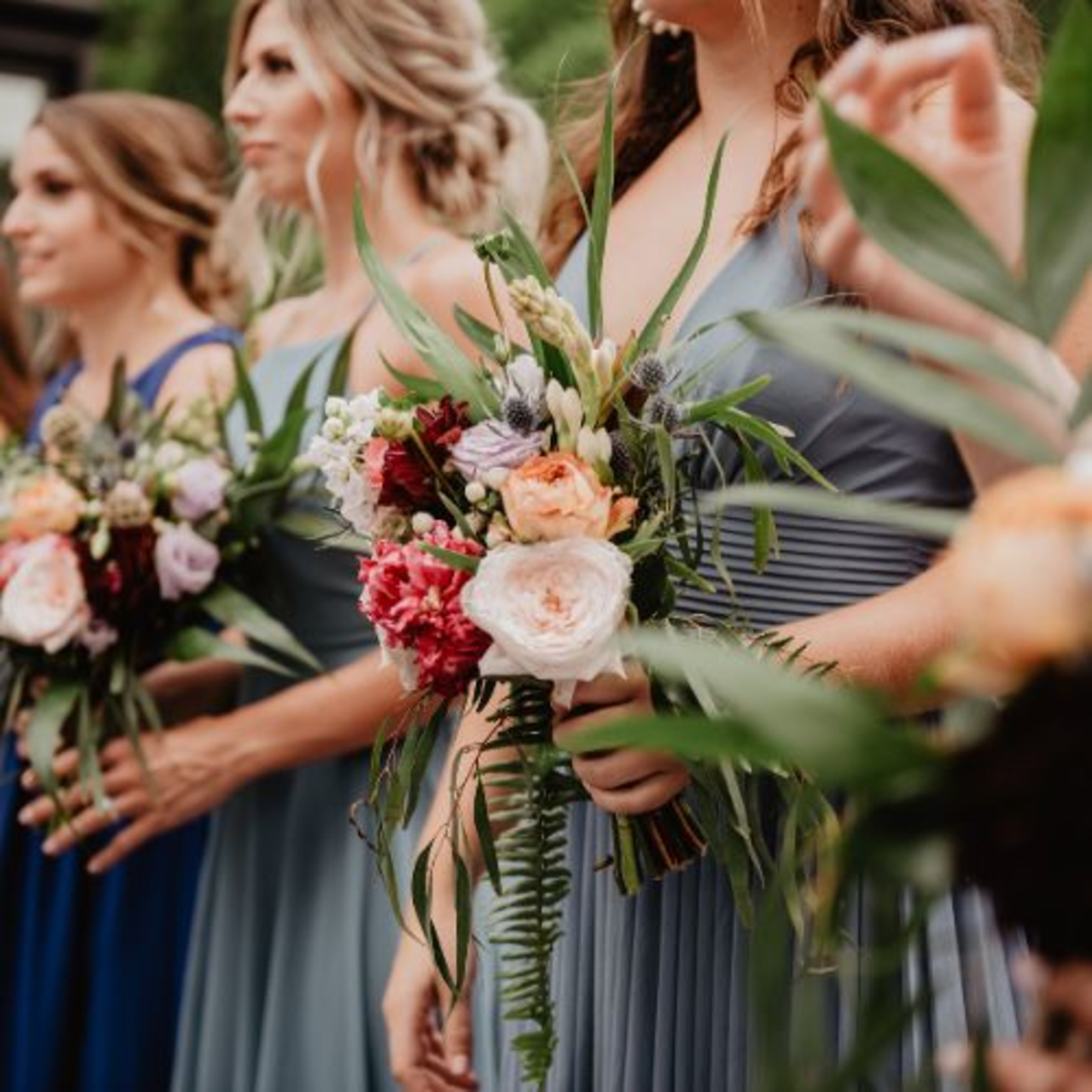 Bridesmaids in blue dresses hold colorful garden-style bouquets at an outdoor wedding.