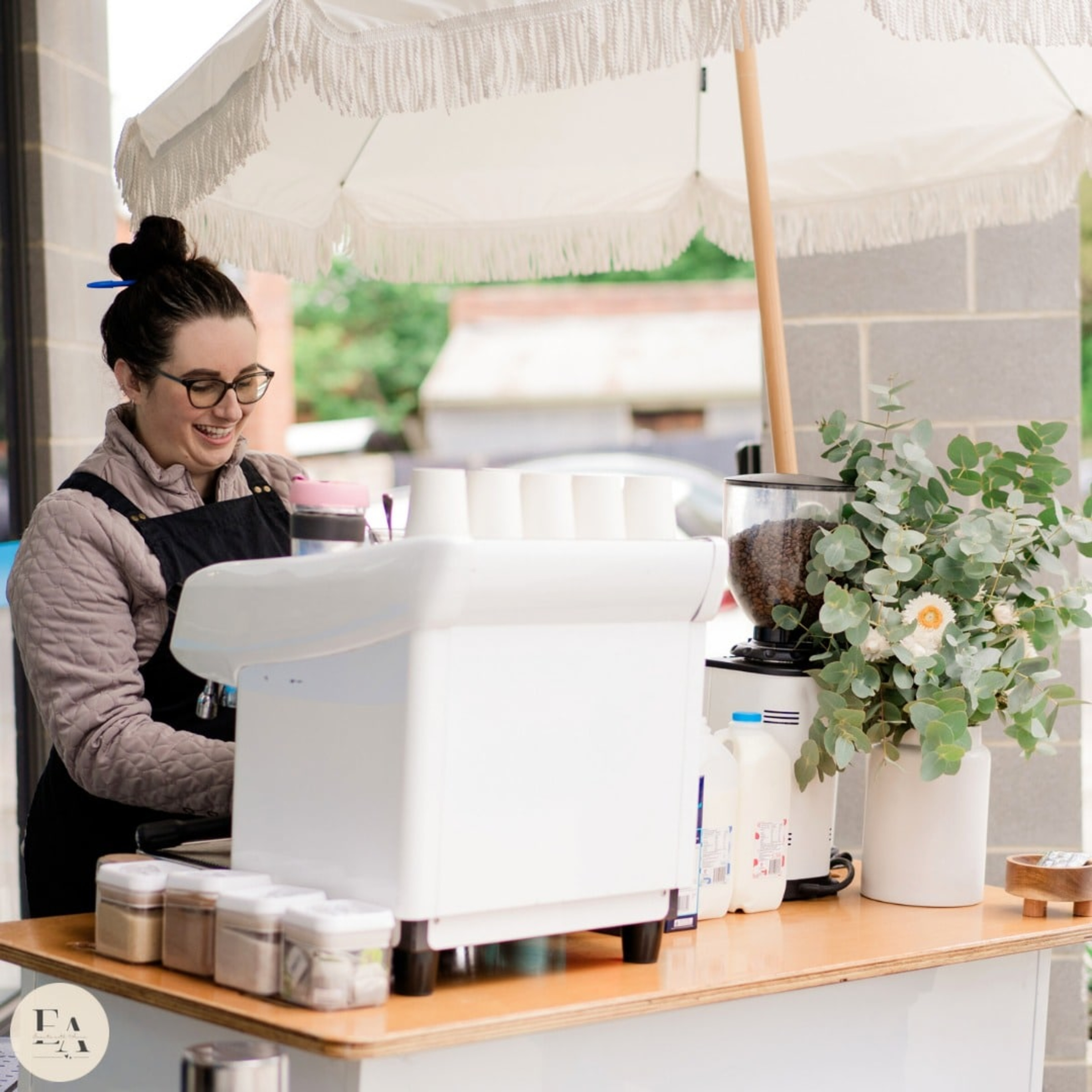 Smiling barista serves coffee from a white mobile espresso cart with greenery under a fringed umbrella.