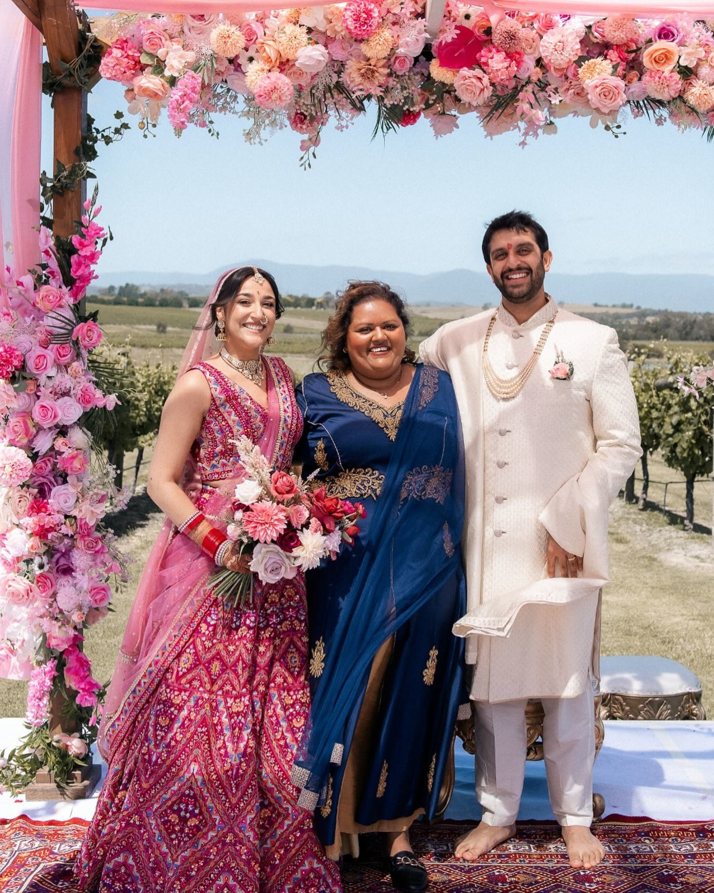 Smiling wedding couple and officiant stand under a vibrant pink floral mandap in a sunny vineyard setting.