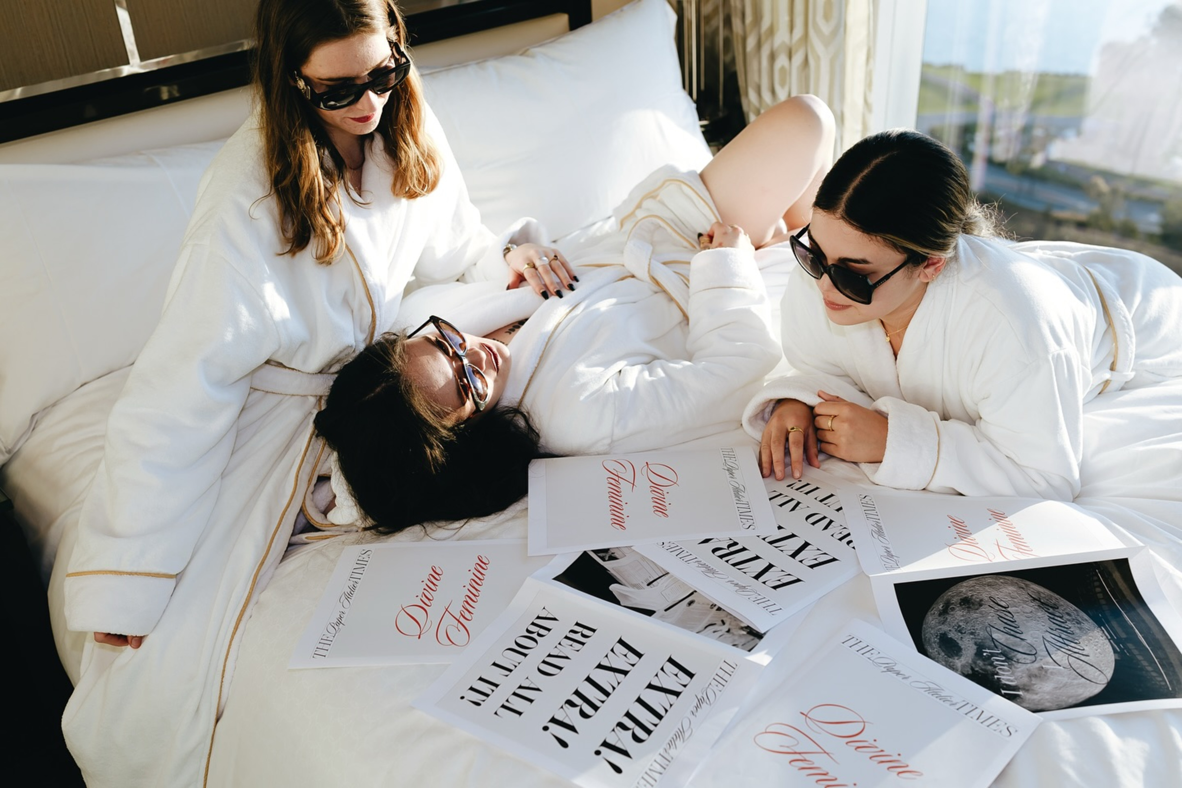 Three women in white robes and sunglasses relax on a hotel bed reviewing stylish wedding magazine layouts.