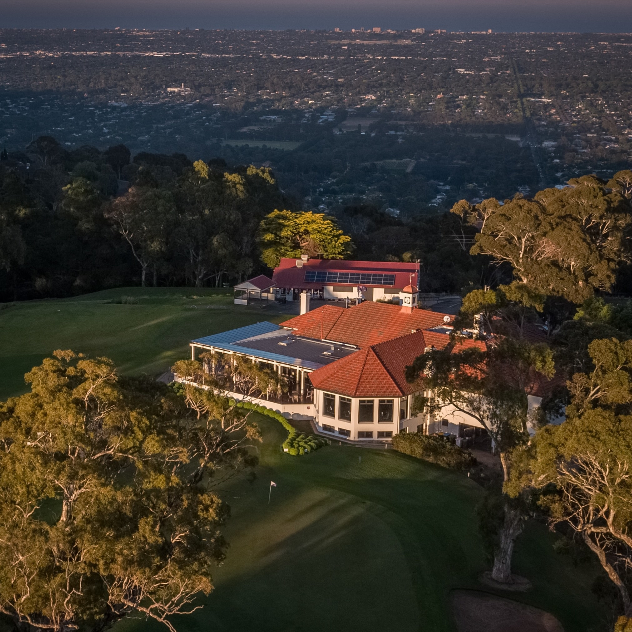A hilltop golf club wedding venue with red-roofed buildings overlooking a city and lush green fairways at sunset.
